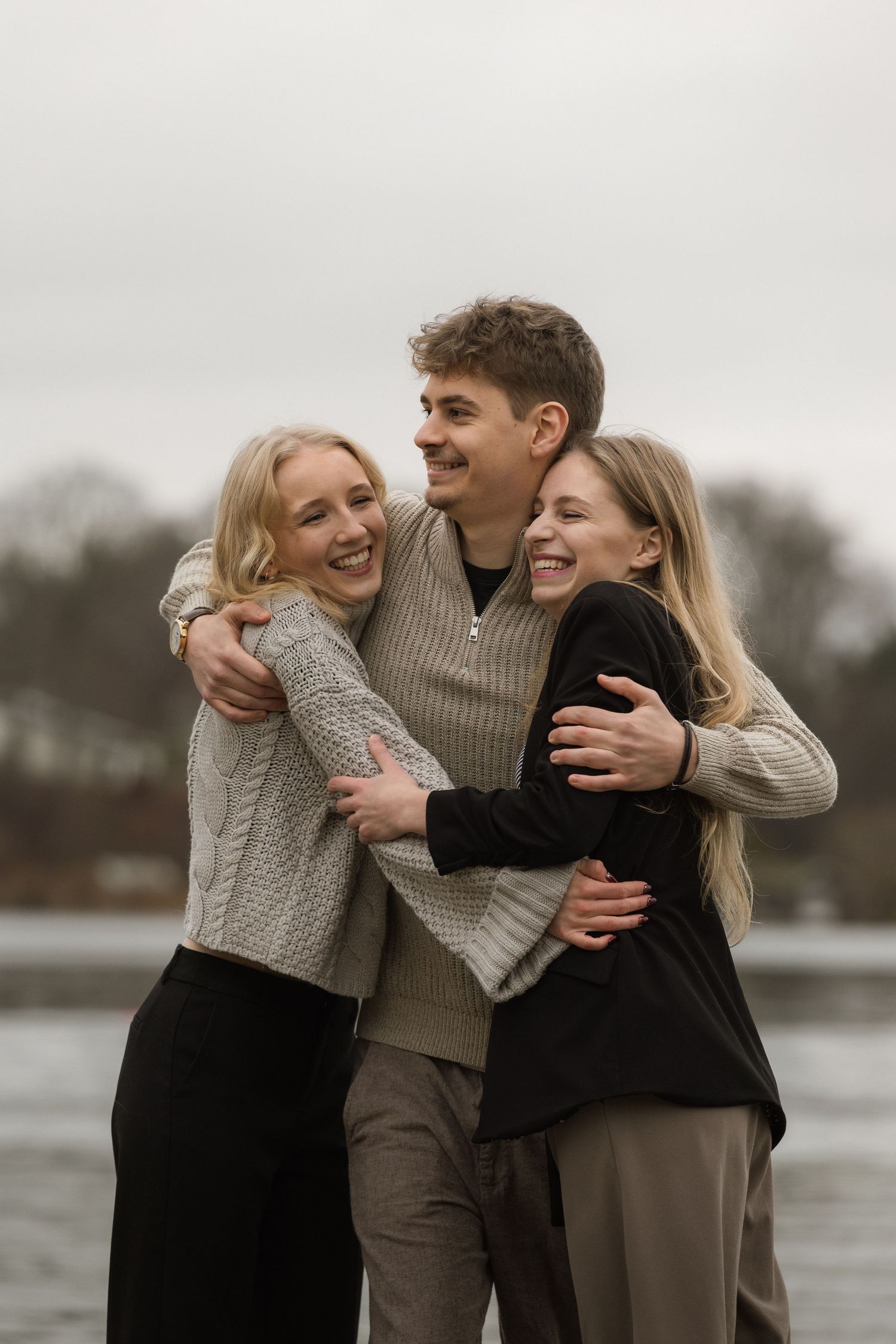 Three laughing young adults share a warm group hug outdoors near a lake in autumn.