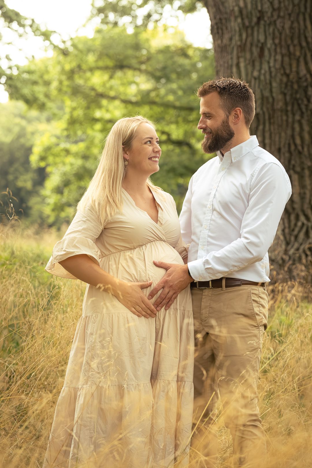 Smiling pregnant couple embracing in golden field, woman in cream dress, man in white shirt.