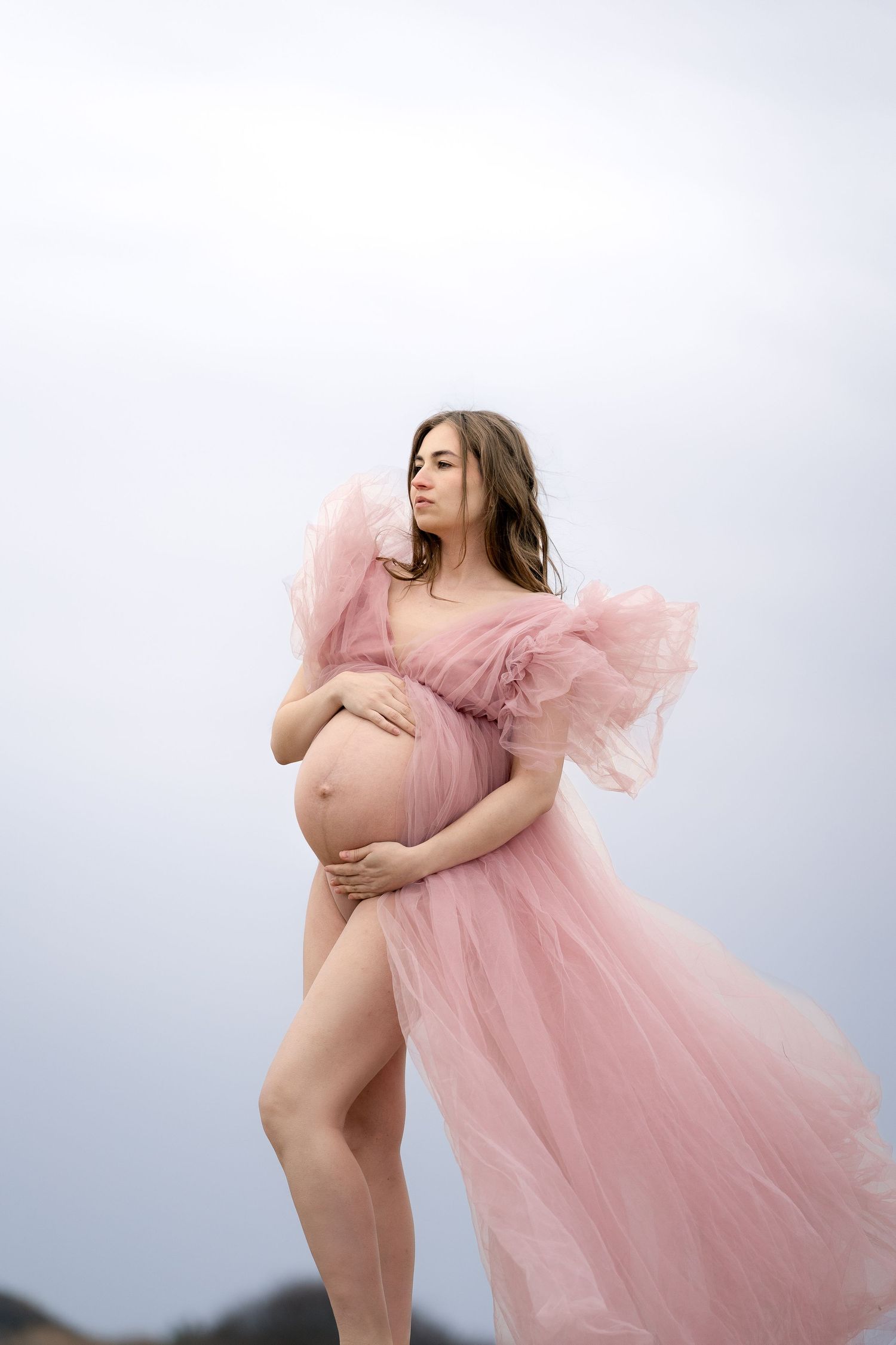 Pregnant woman in pink ruffled tulle maternity gown cradling bump against overcast sky.
