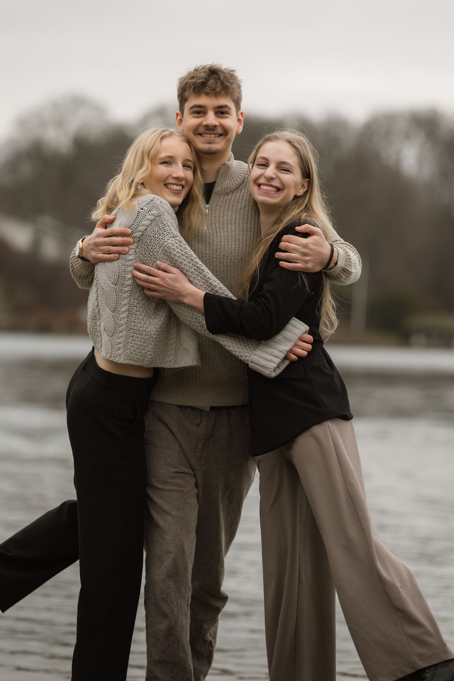 Three joyful young adults embrace and smile together by a lakeside on a cloudy day.