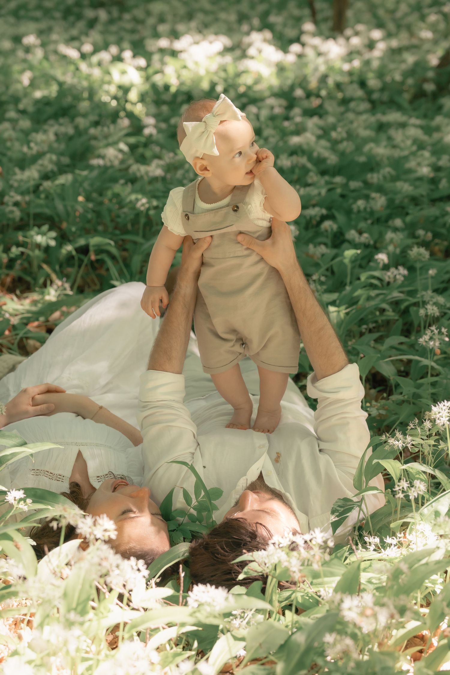 Parents lying in field of white wildflowers while baby sits on fathers chest in forest.