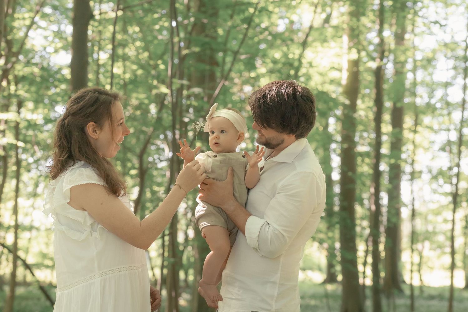 Young couple in white clothing interacting with their baby in a sunlit forest.