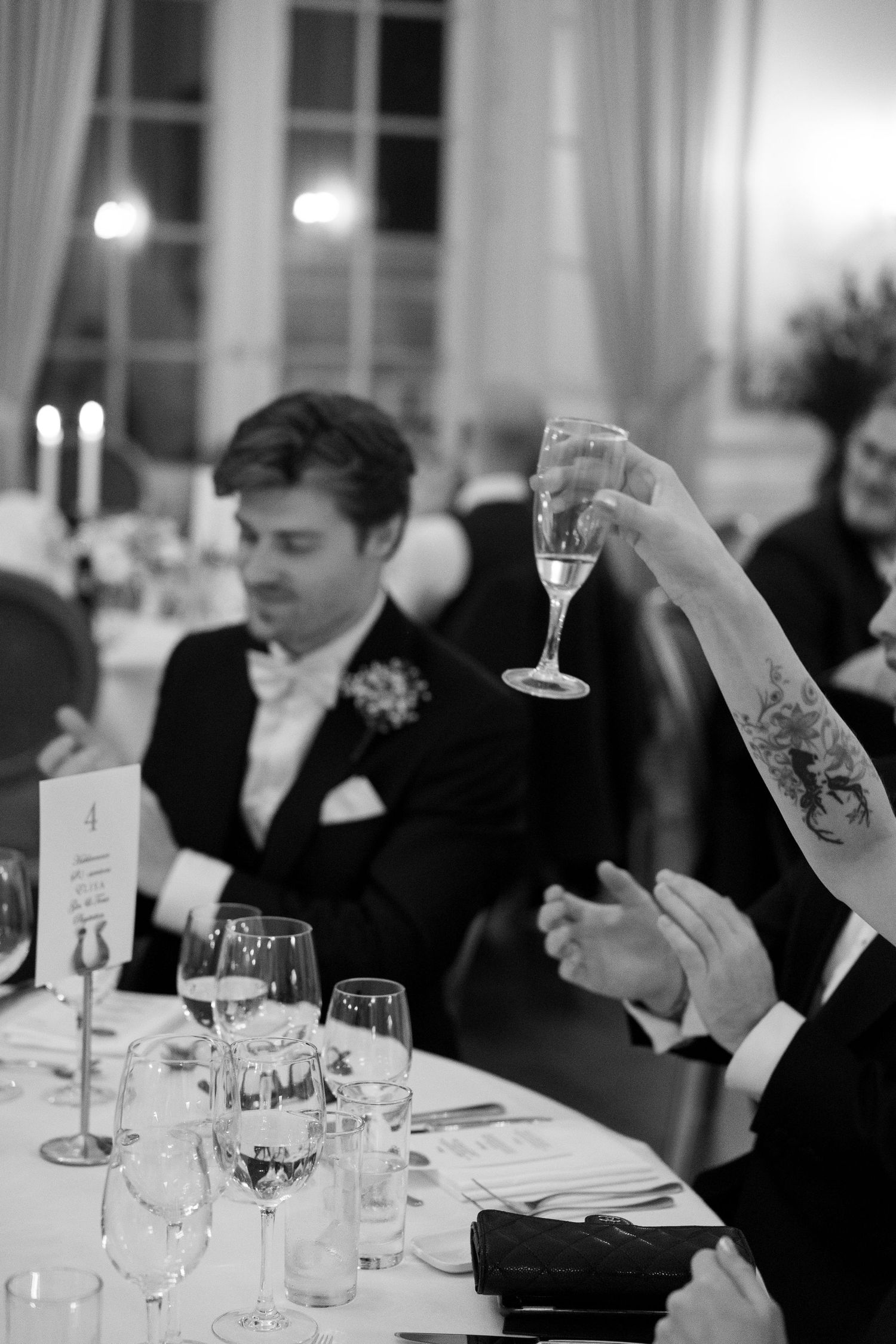 Guest raising champagne glass in toast at formal wedding reception dinner with tattooed arm visible.