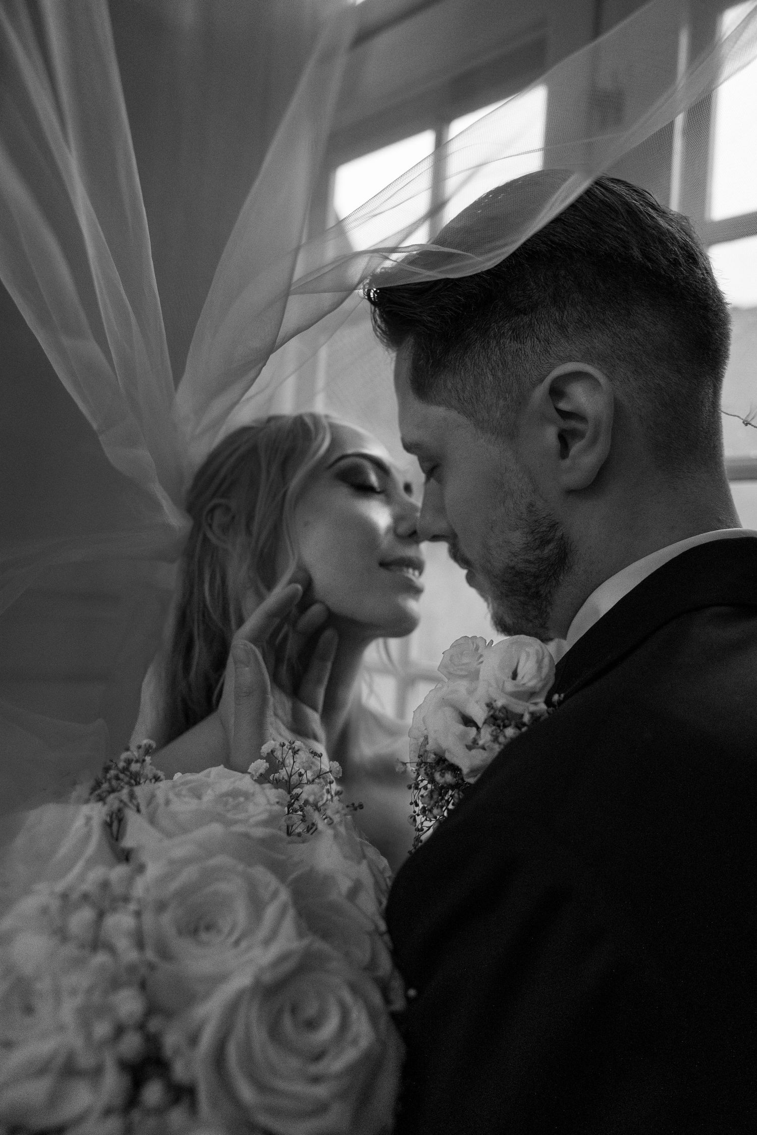 Bride and groom share romantic nose-to-nose moment beneath flowing veil with rose bouquet.