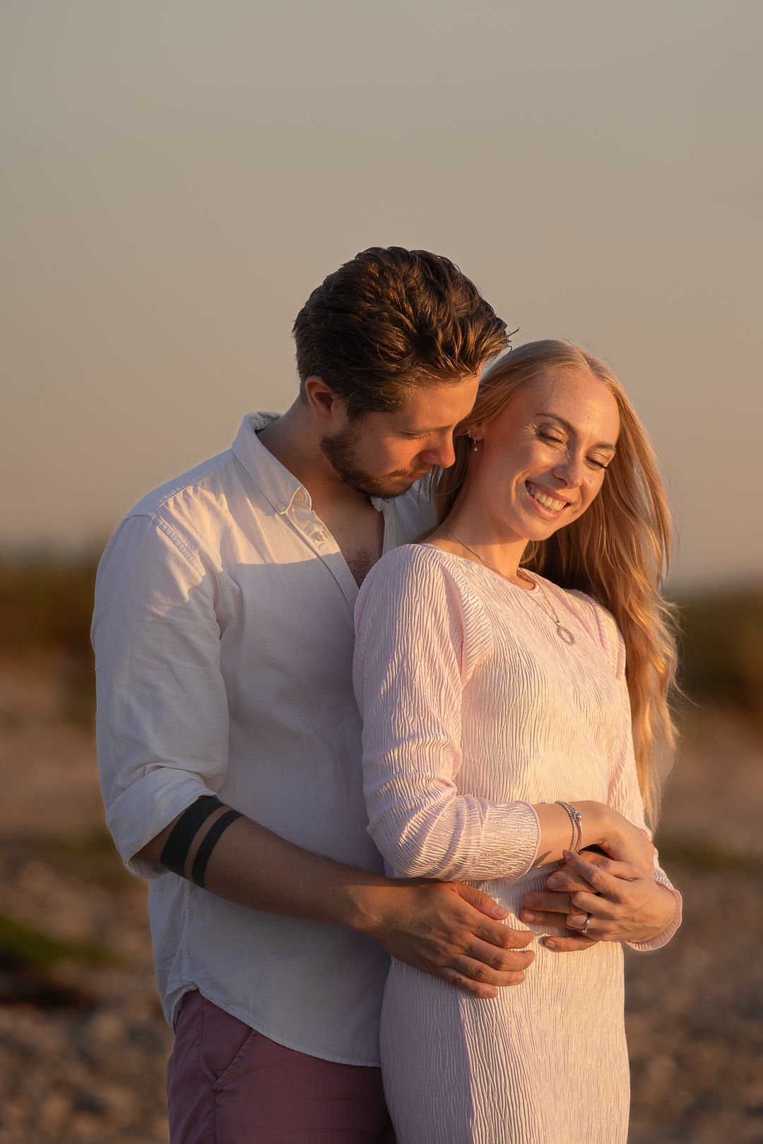Happy couple cuddling on sandy beach at golden sunset, woman smiling joyfully in white dress.