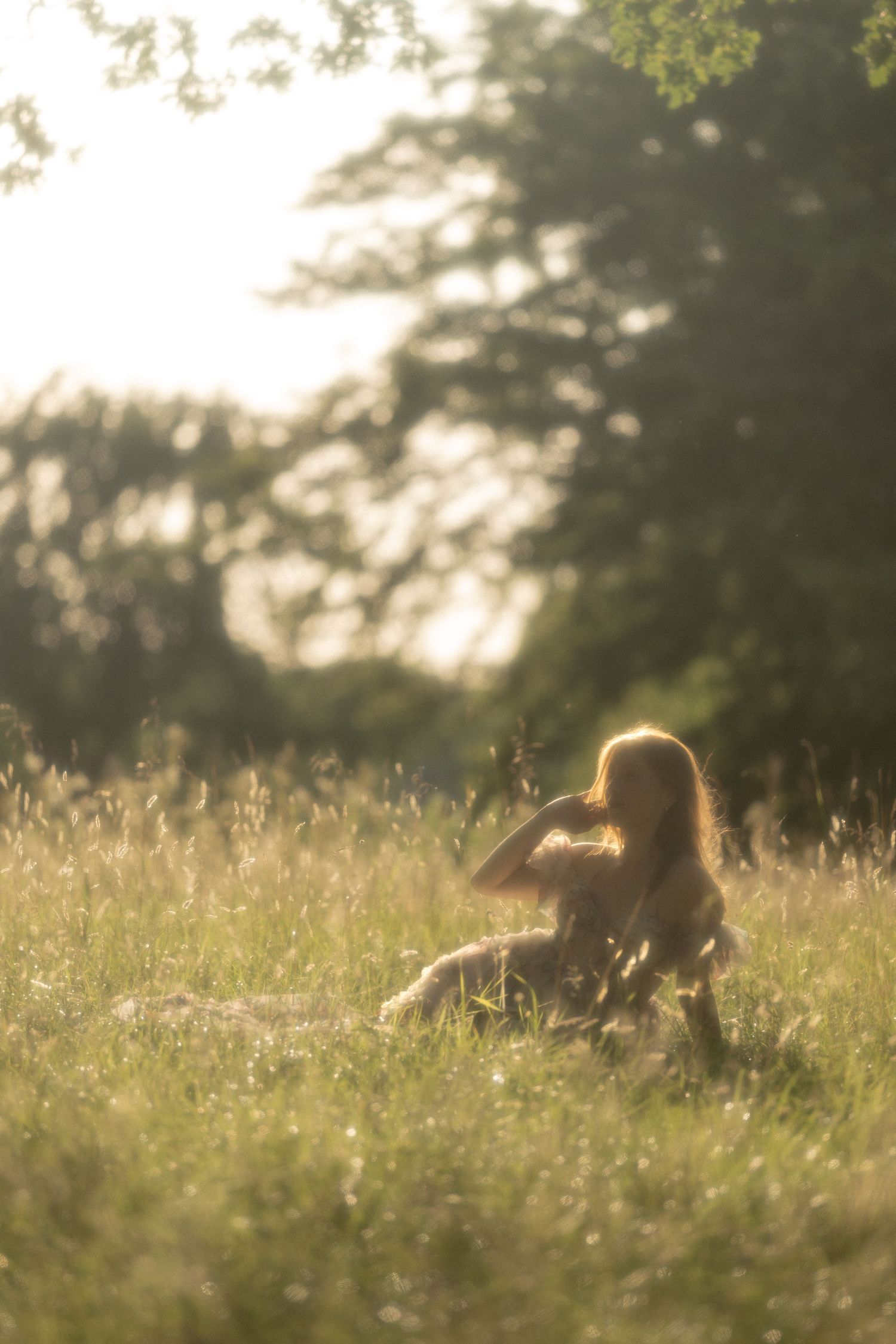 A woman sits peacefully in a sunlit meadow surrounded by tall grass and trees during golden hour.
