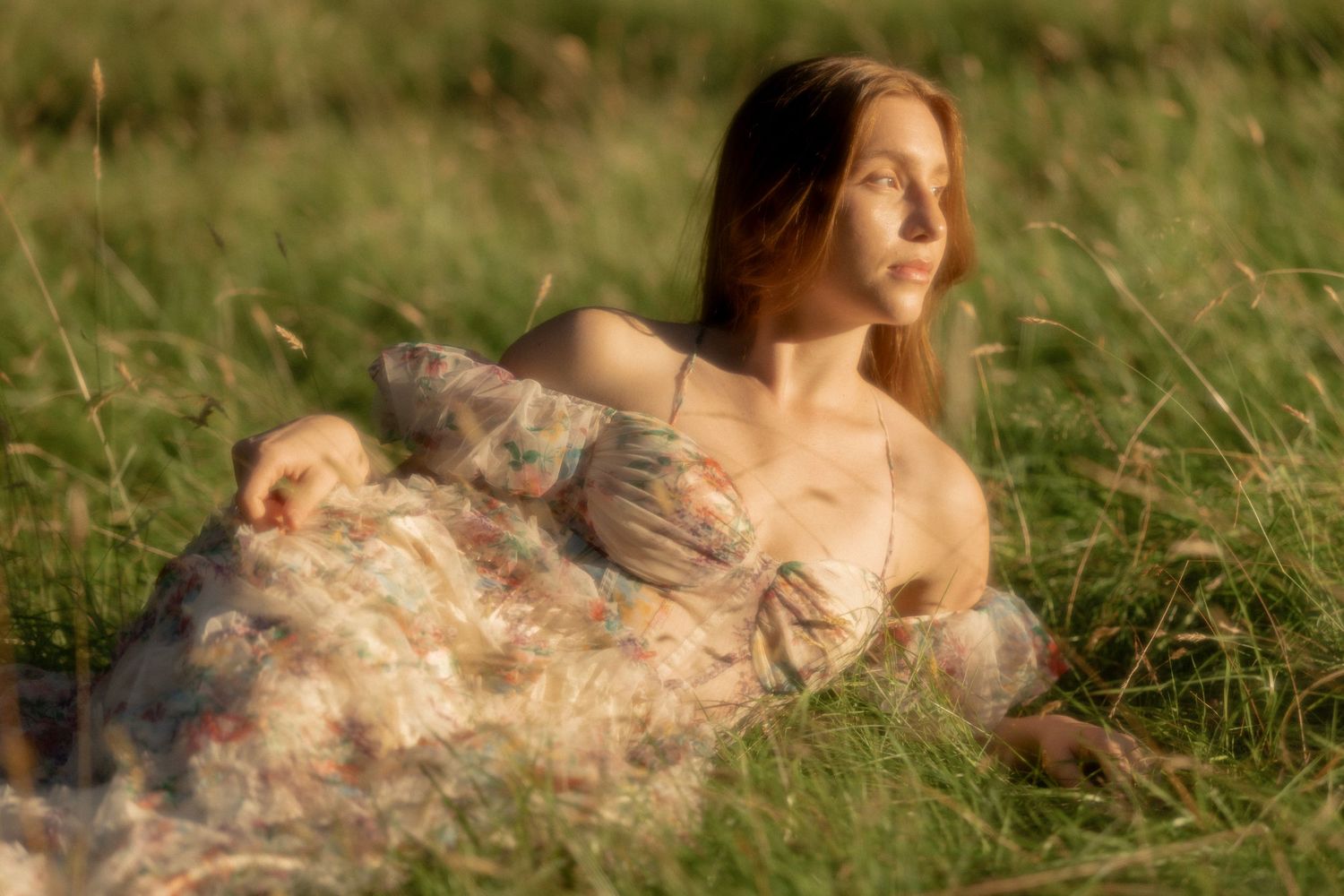 A young woman in a floral dress reclines in a lush green meadow, gazing serenely into the distance.