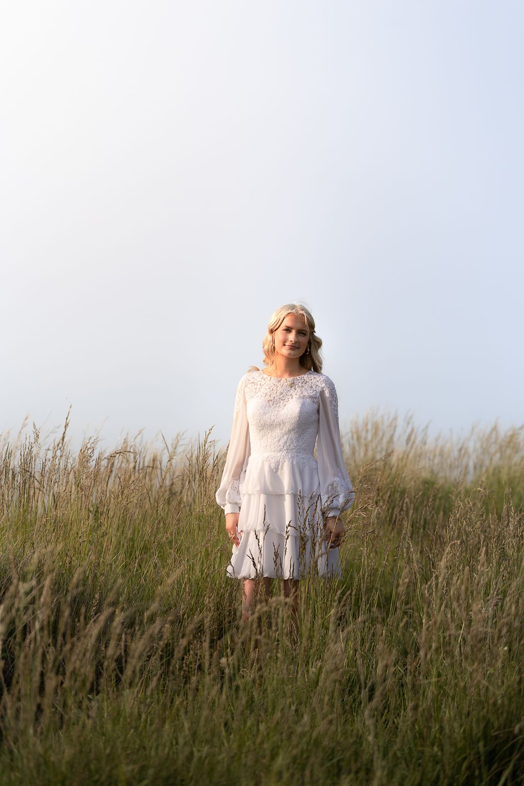 Blonde woman in white tiered lace dress posing in lush green tall grass field.