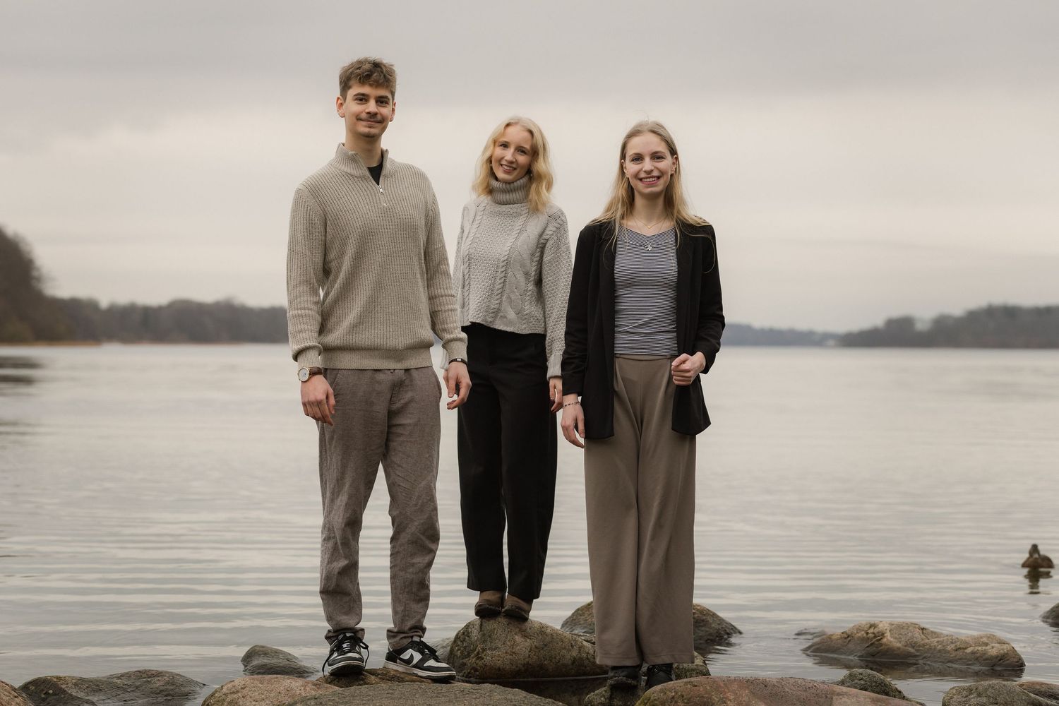 Three smiling young adults stand on rocks by a serene lake under a cloudy sky.