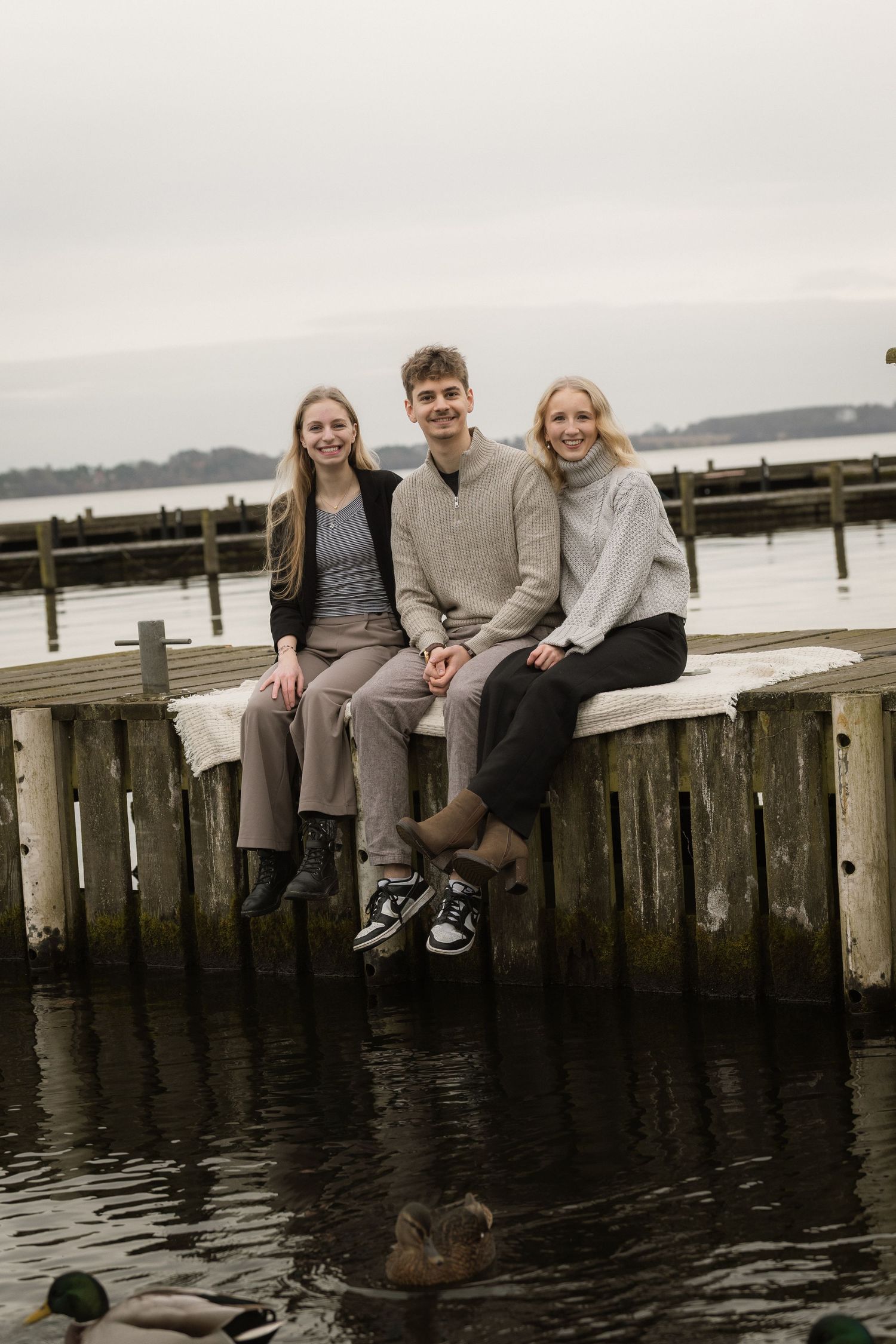 Three smiling young adults sit together on a wooden pier over calm water with a duck below.