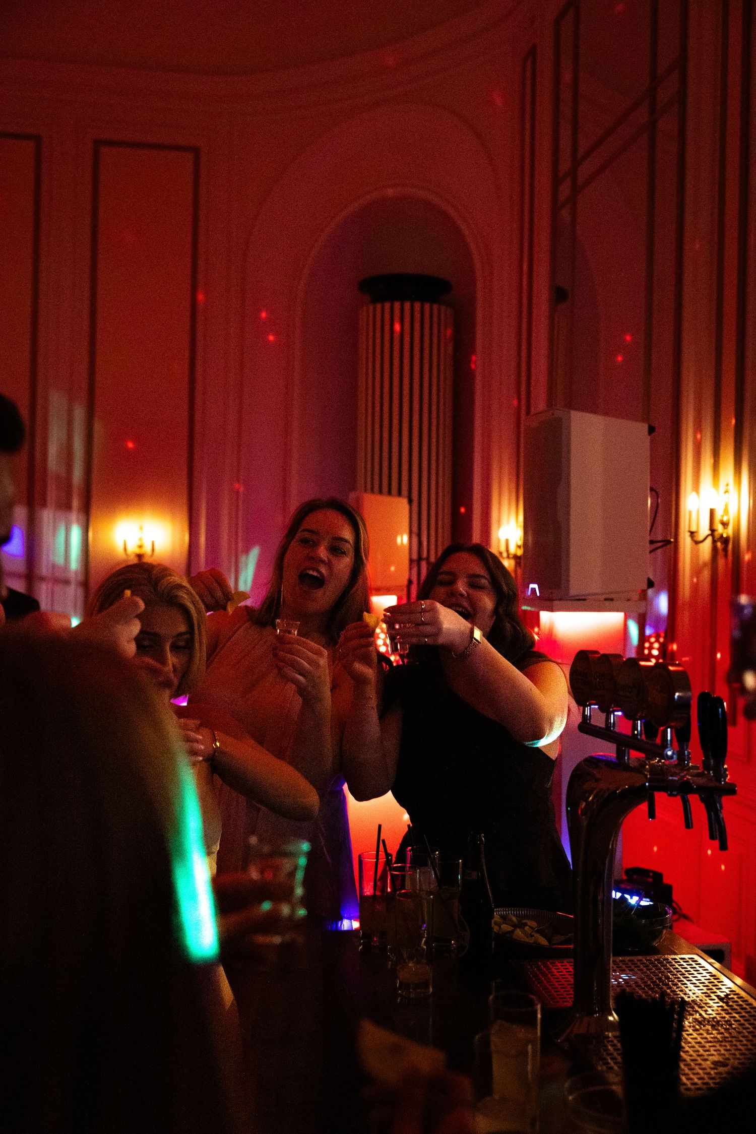 Wedding guests taking shots at a bar bathed in warm red and orange lighting during a lively reception party.