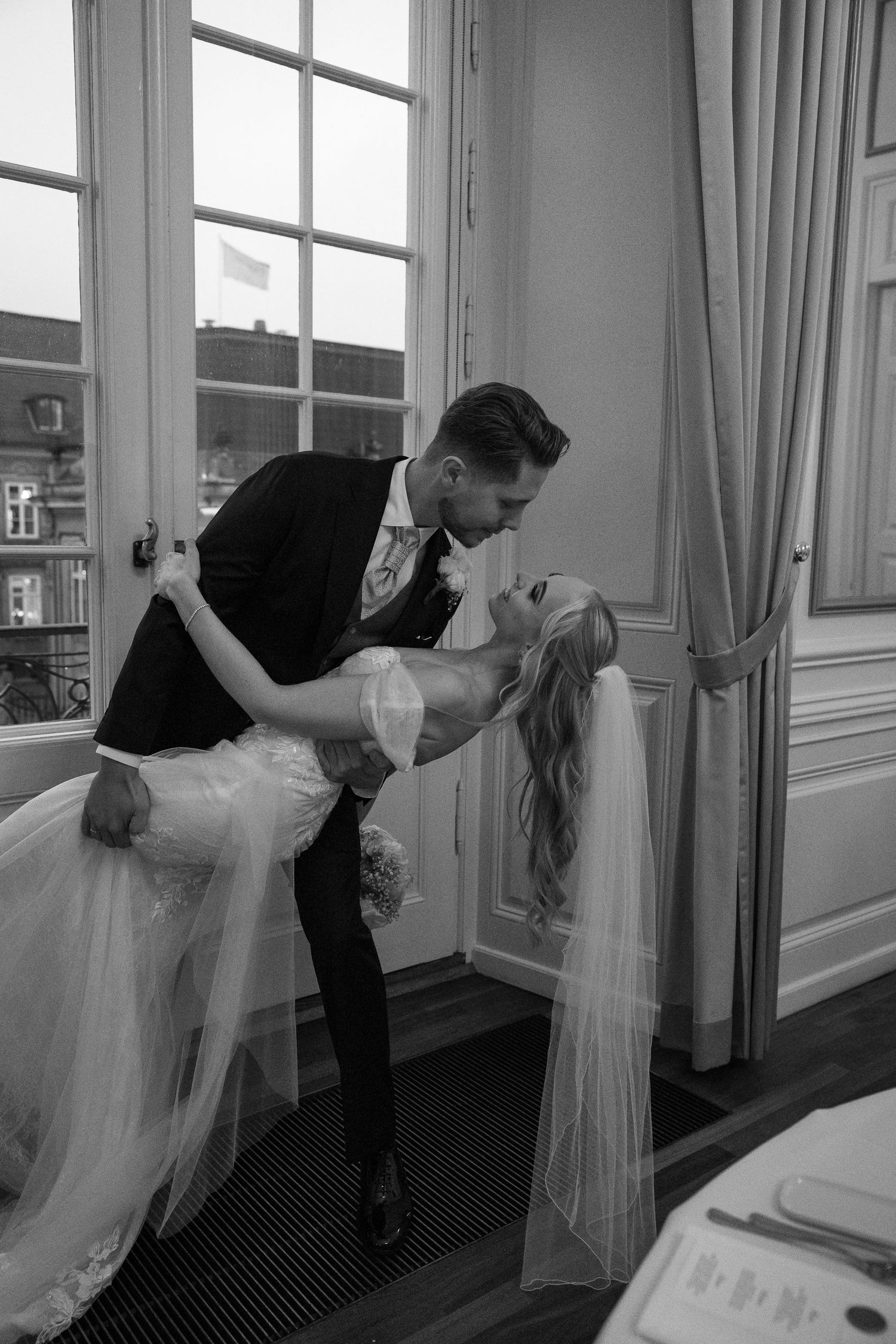 Black and white photo of groom dipping bride for a kiss near elegant French windows at wedding reception.