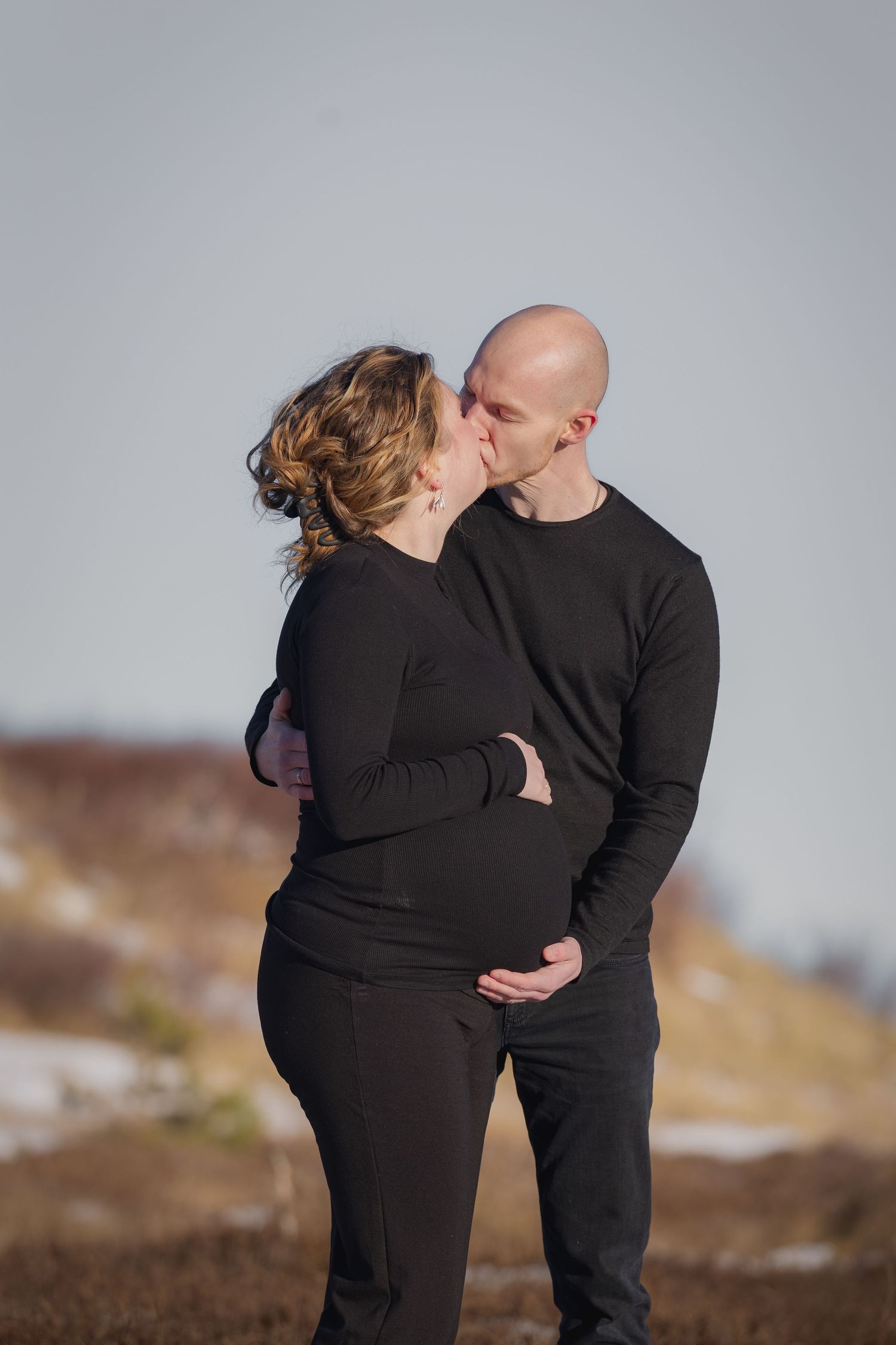 Expecting couple in matching black outfits sharing a kiss outdoors in snowy winter landscape.