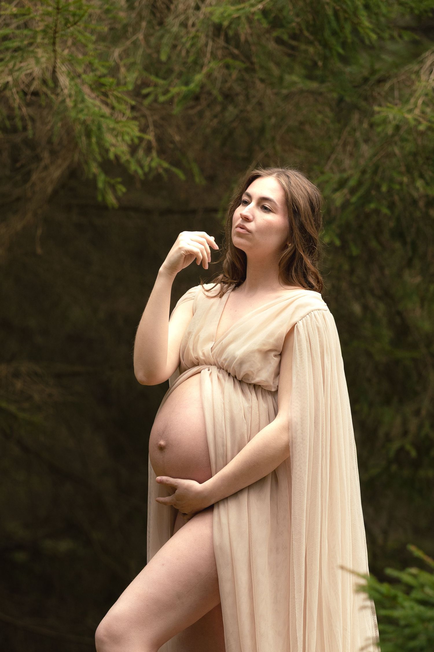 Pregnant woman in sheer beige draped maternity gown posing thoughtfully in a forest setting.