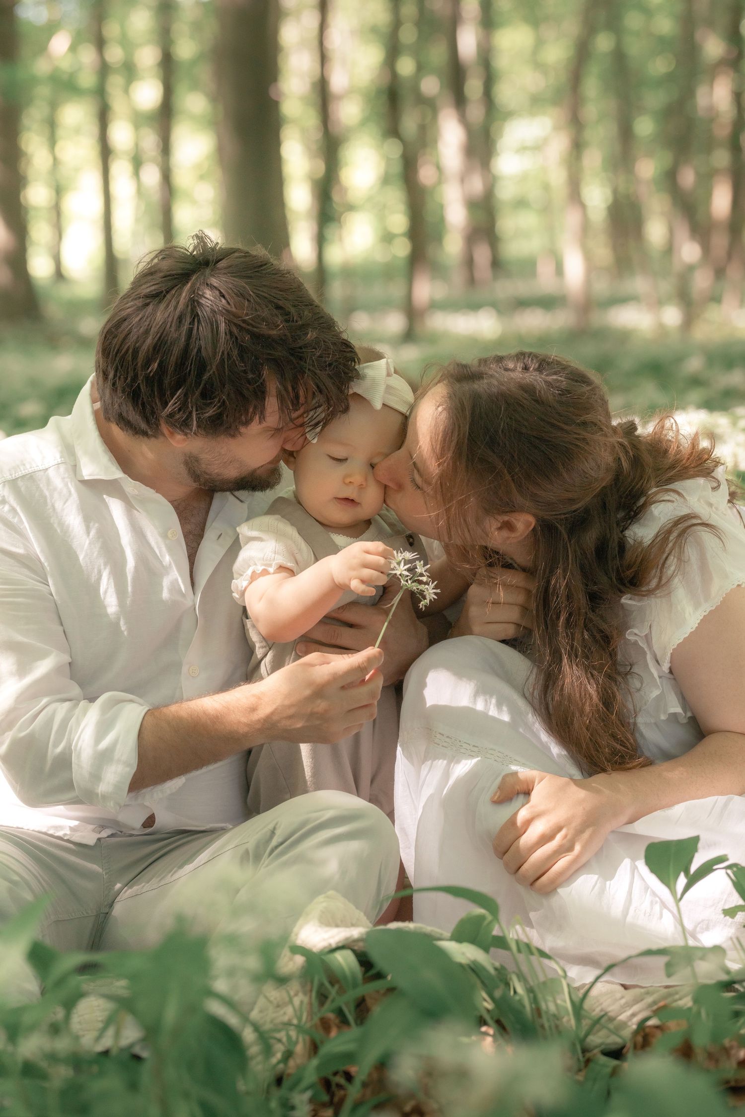 Parents share a kiss with their baby while sitting among wildflowers in a sunny forest.