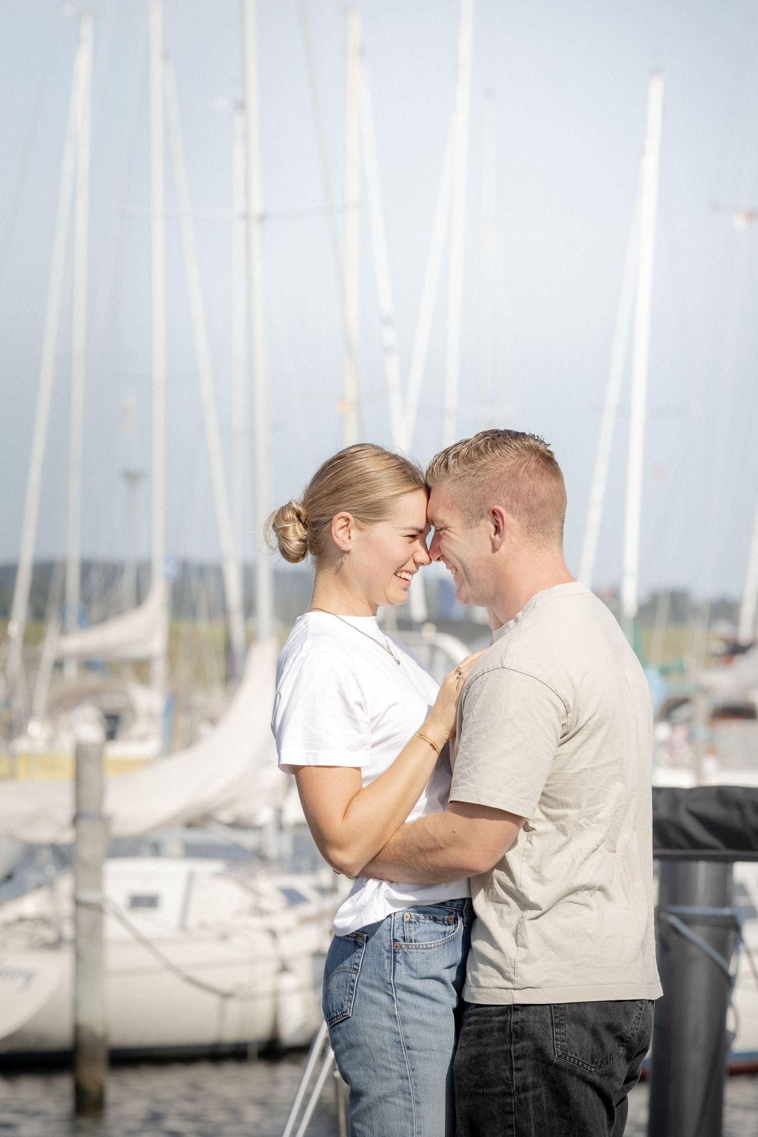 Romantic couple touching foreheads and smiling at a sunny marina with sailboats behind them.