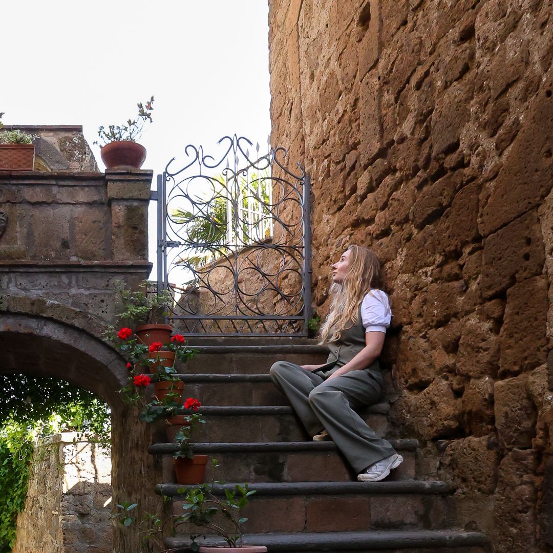 A young blonde woman sits on stone steps beside a rustic wall with a decorative iron gate and red roses.
