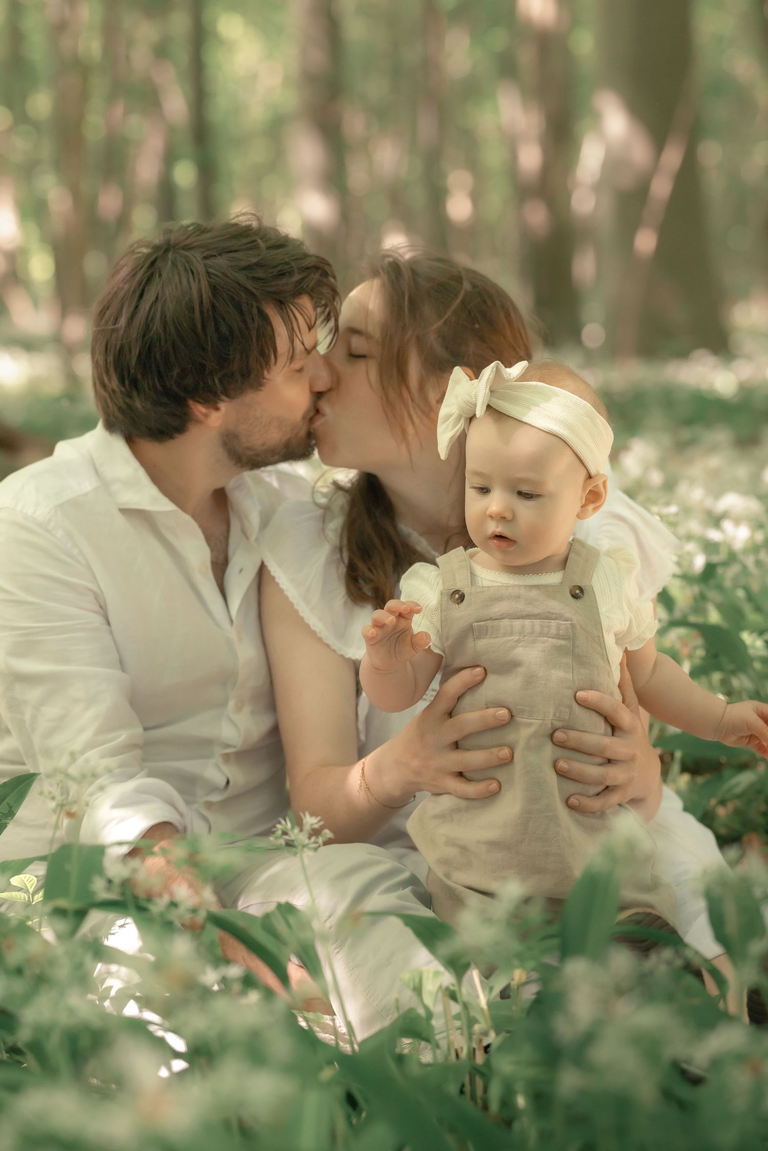 Parents kissing while holding their baby in white overalls among wildflowers in a forest.