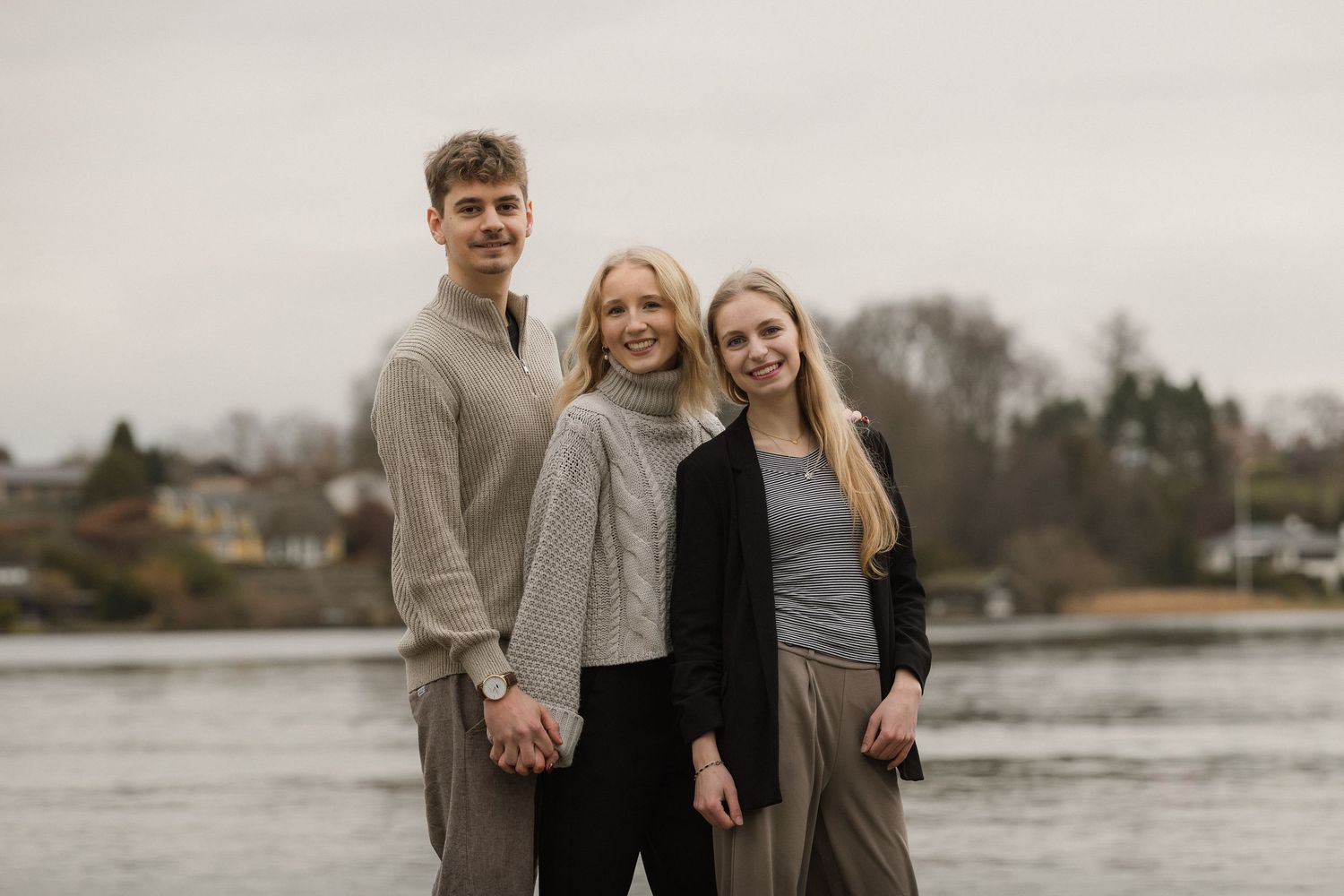 Three smiling teenagers standing together near a river on an overcast day.
