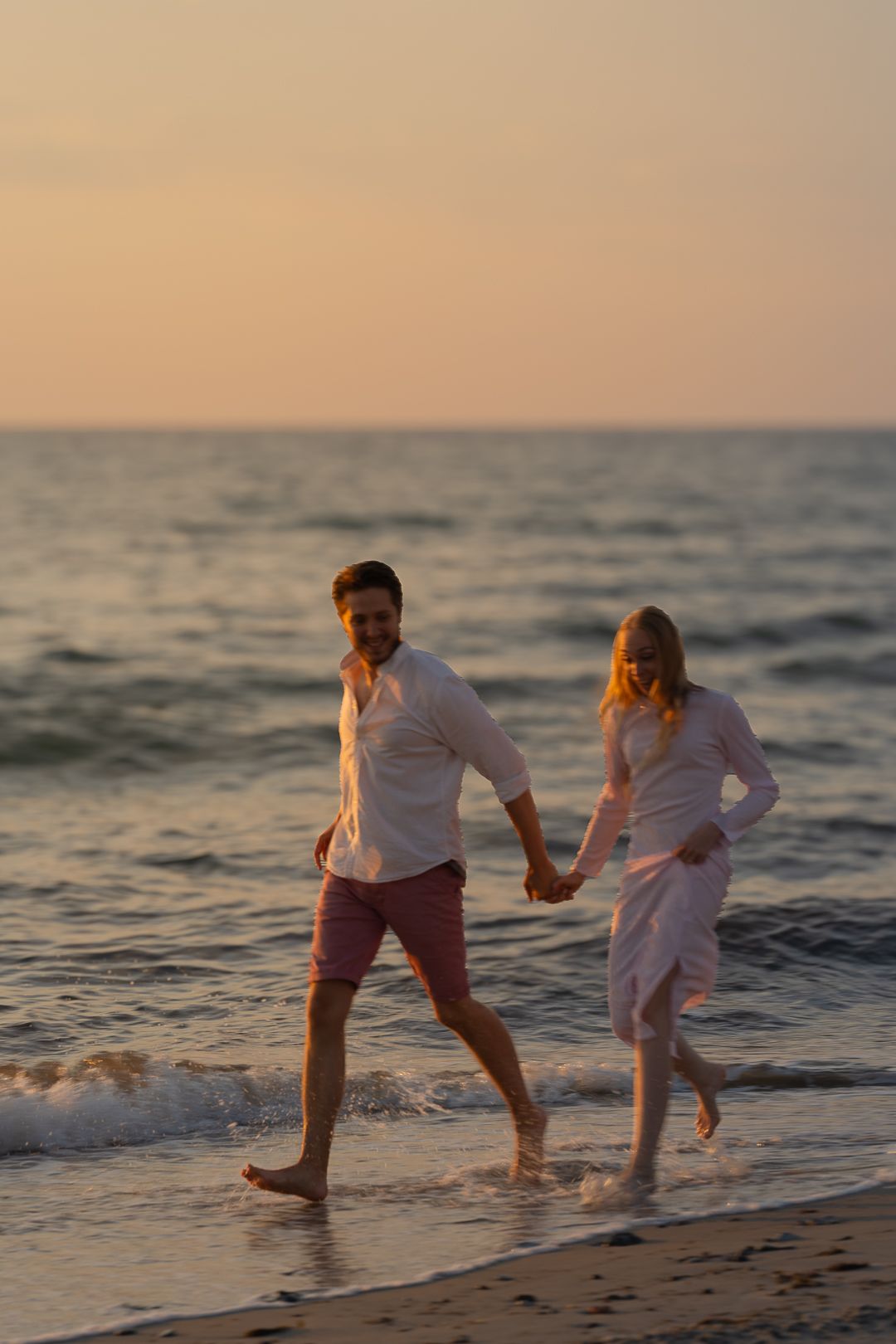 A couple holds hands and runs barefoot along a sandy beach shore during a beautiful golden sunset.