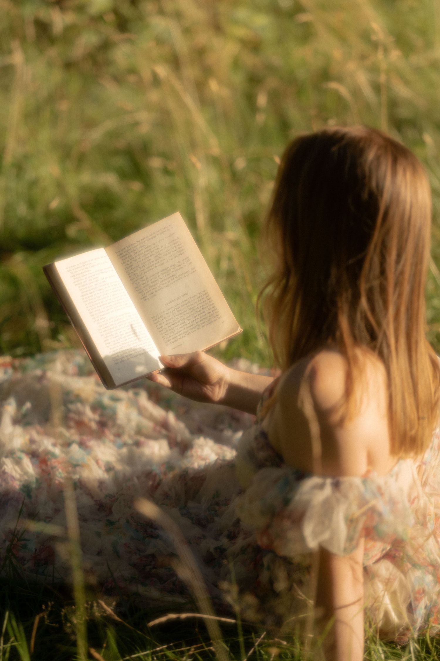 A young girl in a floral dress sits in a sunlit meadow, reading an open book outdoors.