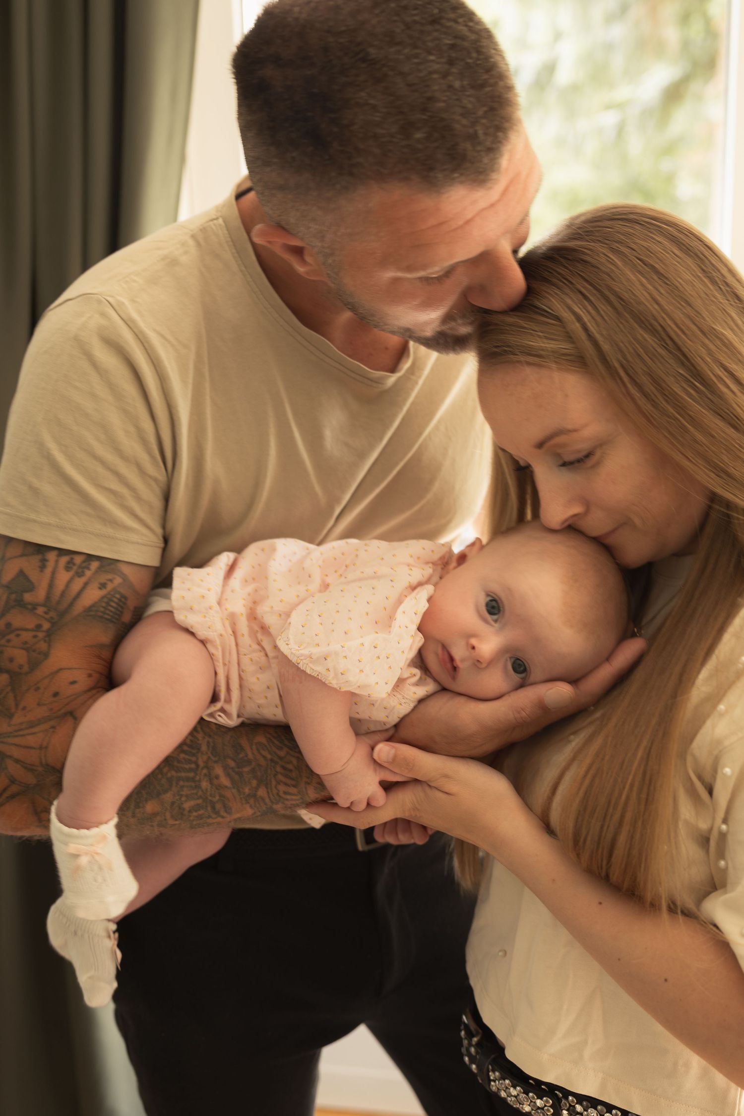 A tattooed father and blonde mother tenderly kiss their newborn baby girl in a bright, warm indoor setting.