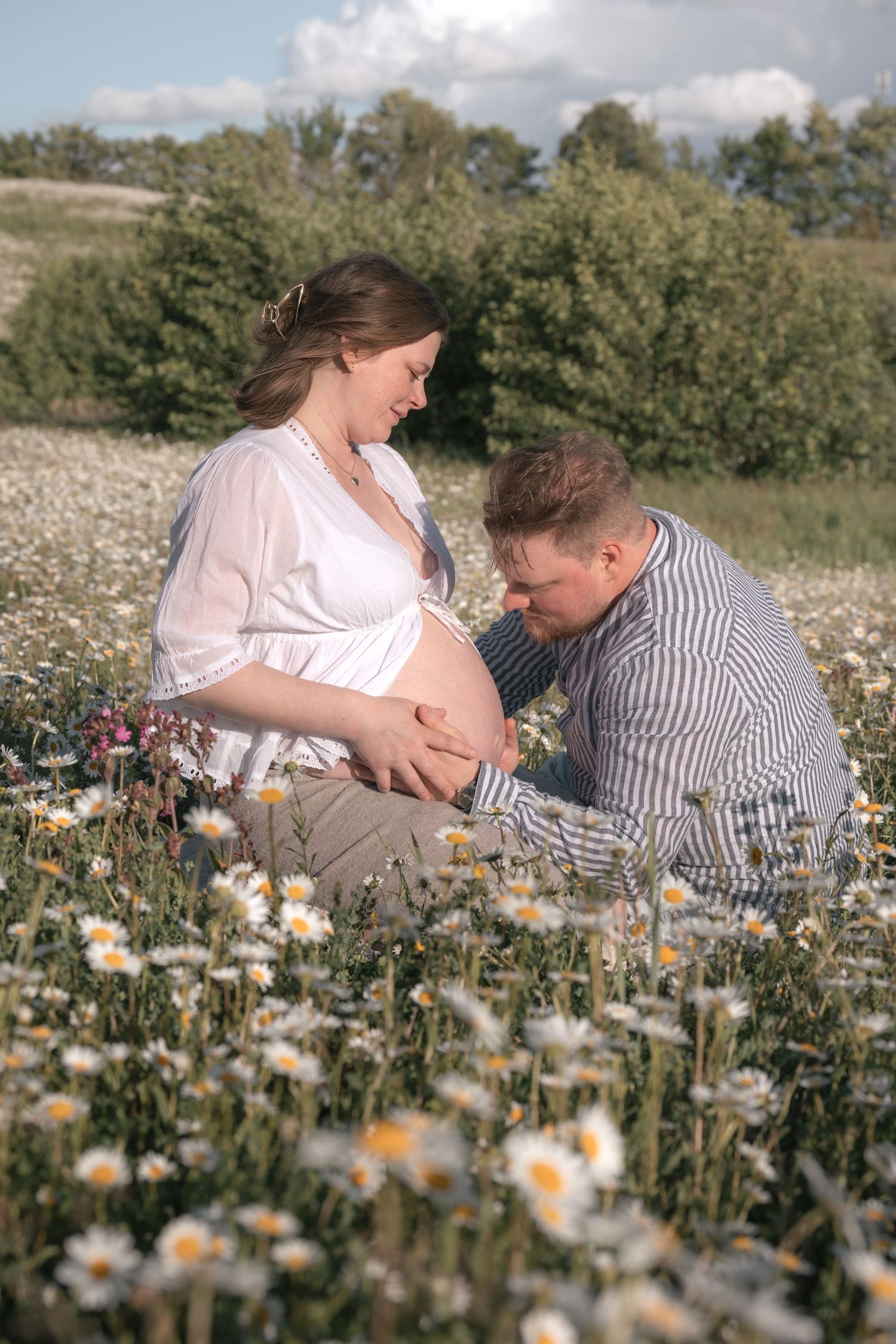 Couple sitting in a daisy field, man kissing pregnant woman's baby bump on a sunny day.