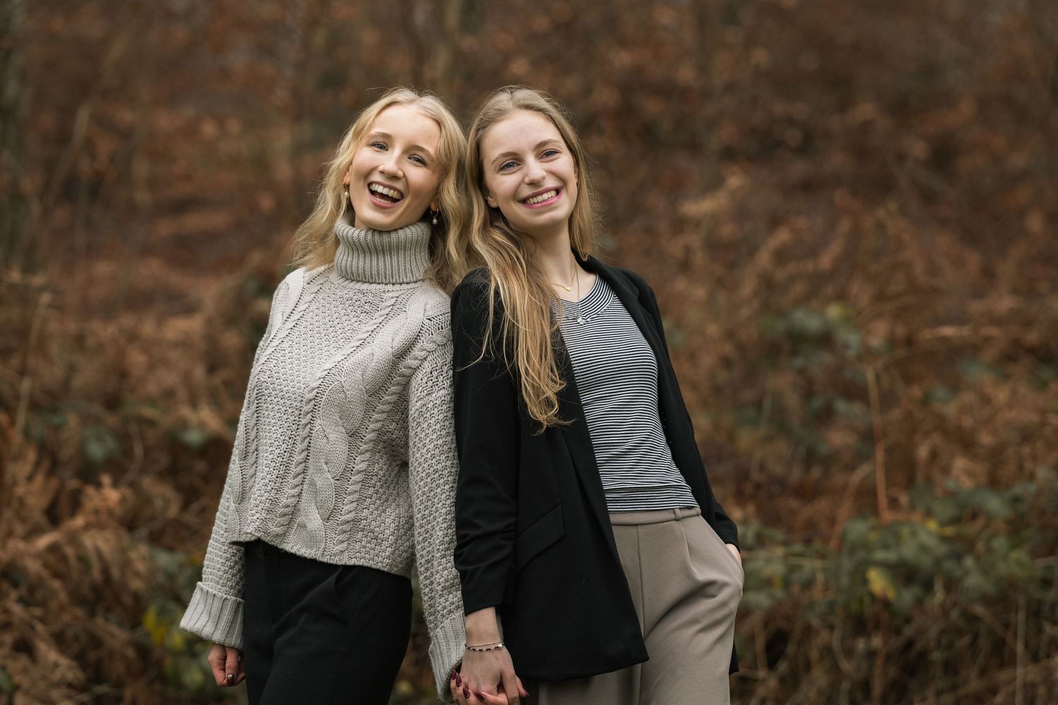 Two smiling blonde women standing back to back holding hands in an autumn woodland setting.