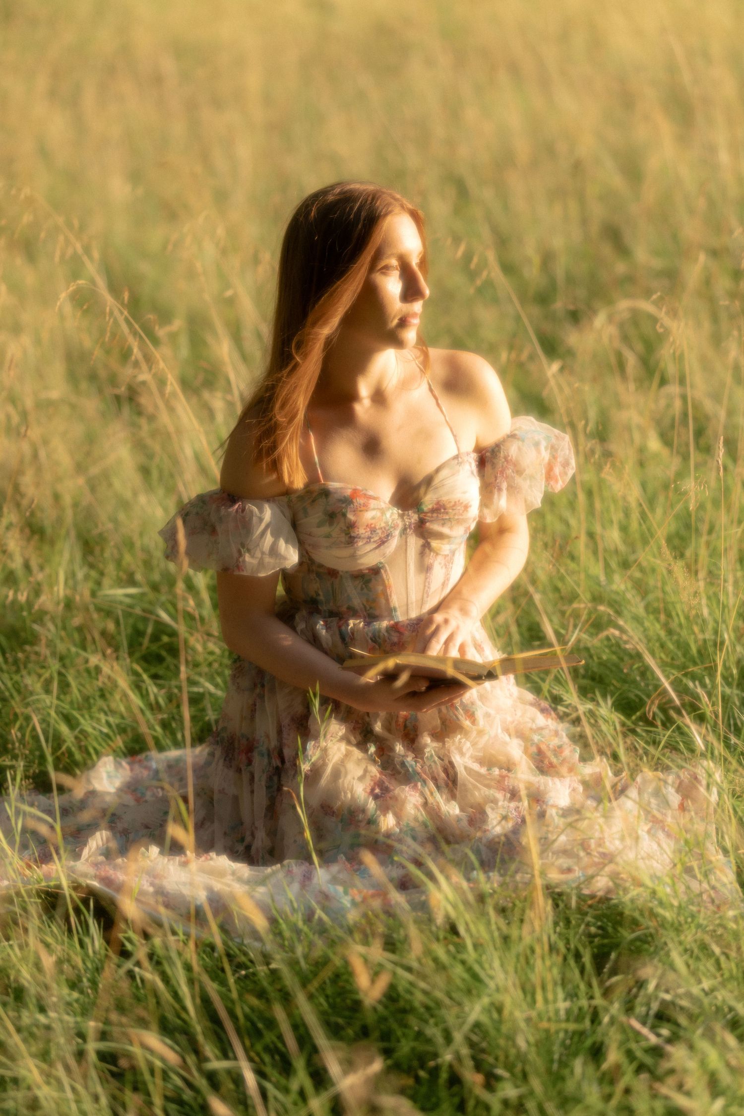 Woman in floral off-shoulder dress sitting gracefully in golden tall grass field during warm sunset light.
