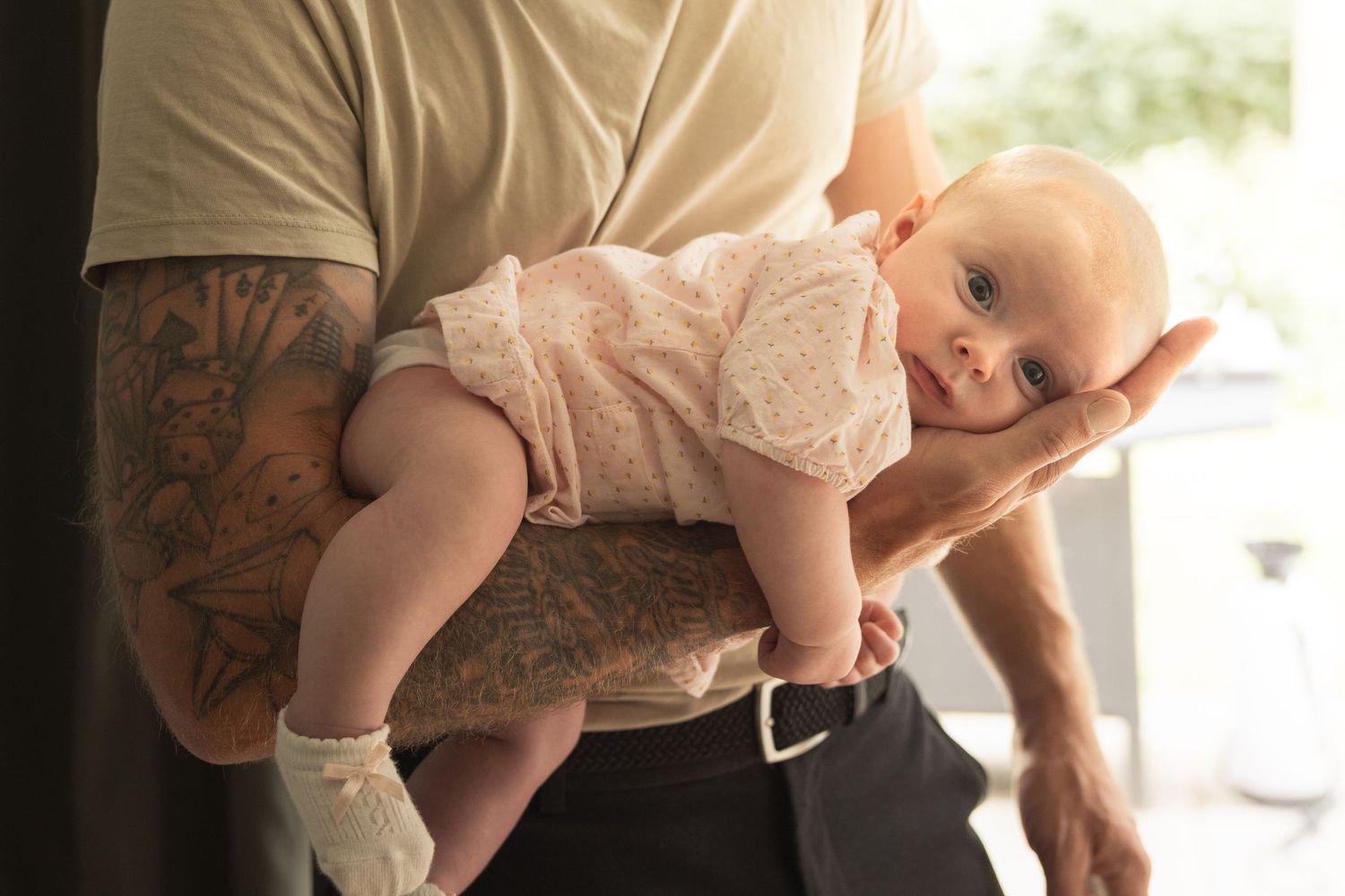 Tattooed father carefully holding his newborn baby in pink onesie, supporting the infant with both arms.