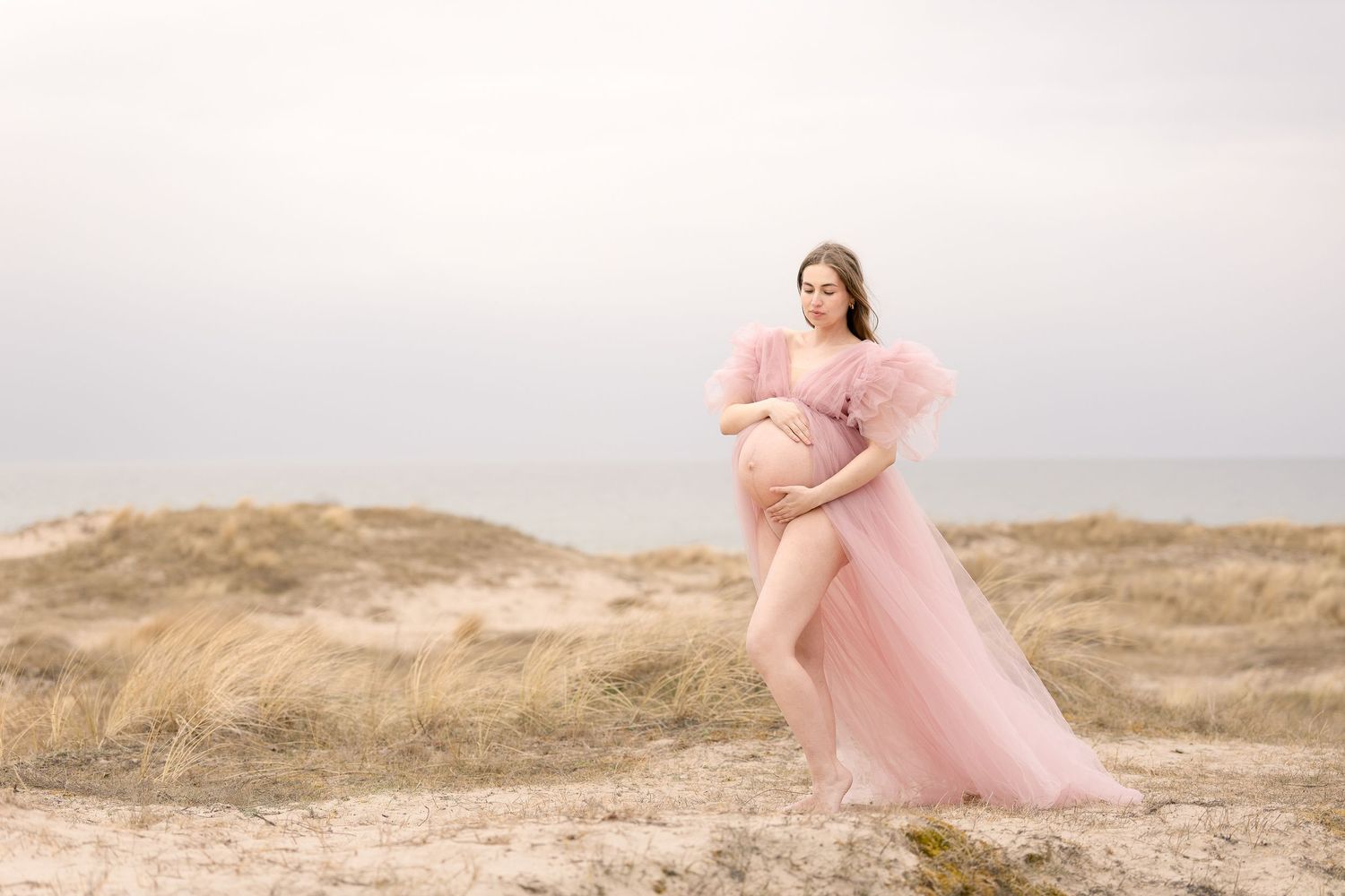 Pregnant woman in pink tulle maternity gown posing on sandy beach dunes with flowing skirt.