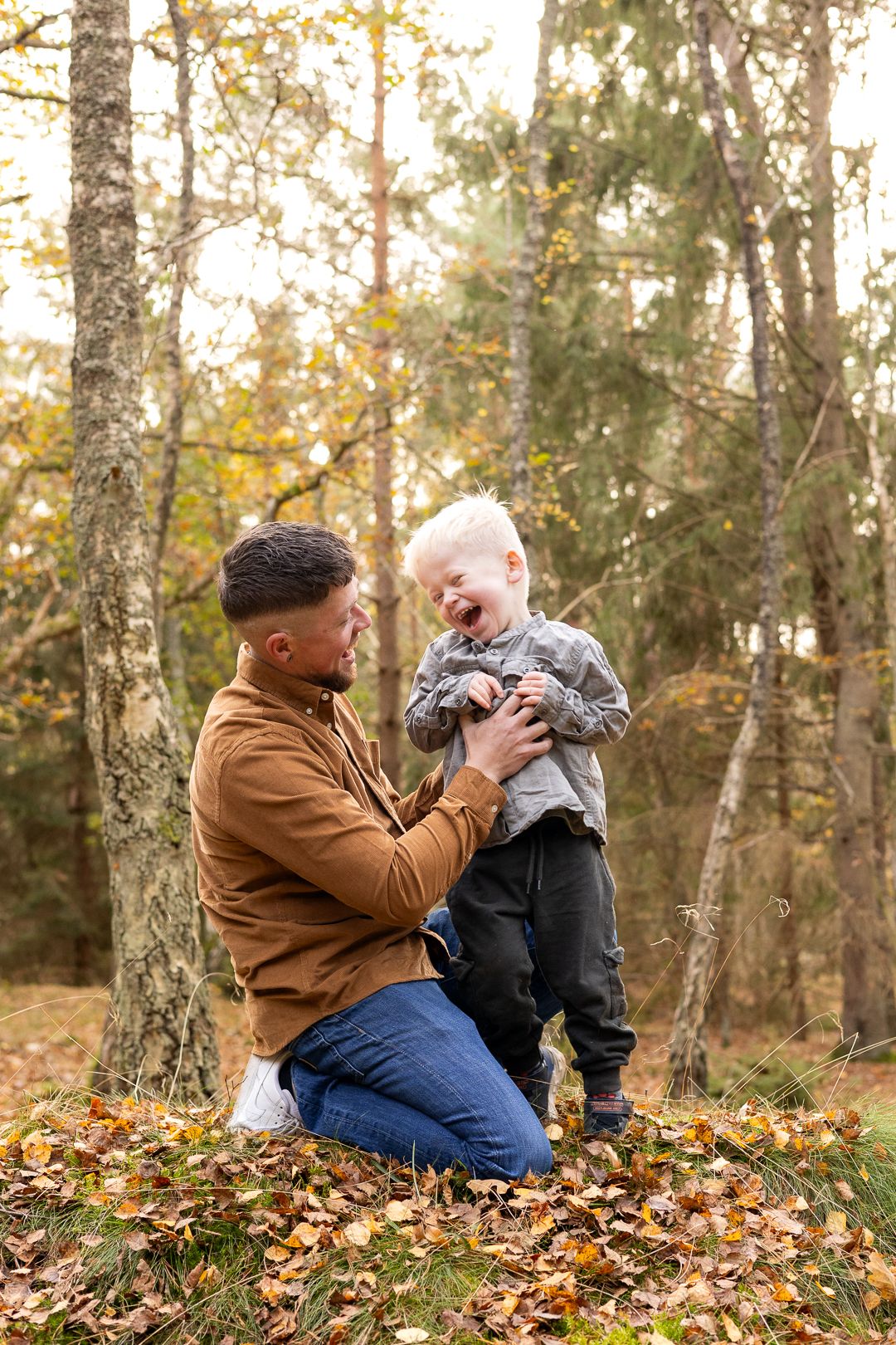 Father kneeling beside his giggling toddler son in a sunlit autumn forest.