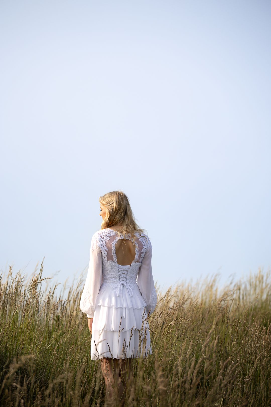 Blonde girl in white lace tiered dress standing in tall grass field under blue sky.