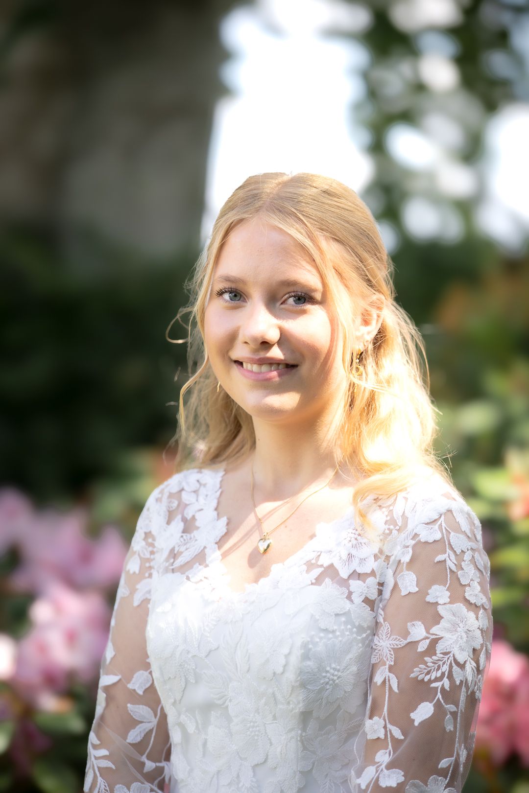 Smiling blonde teen girl in white lace dress posing outdoors among pink flowers.