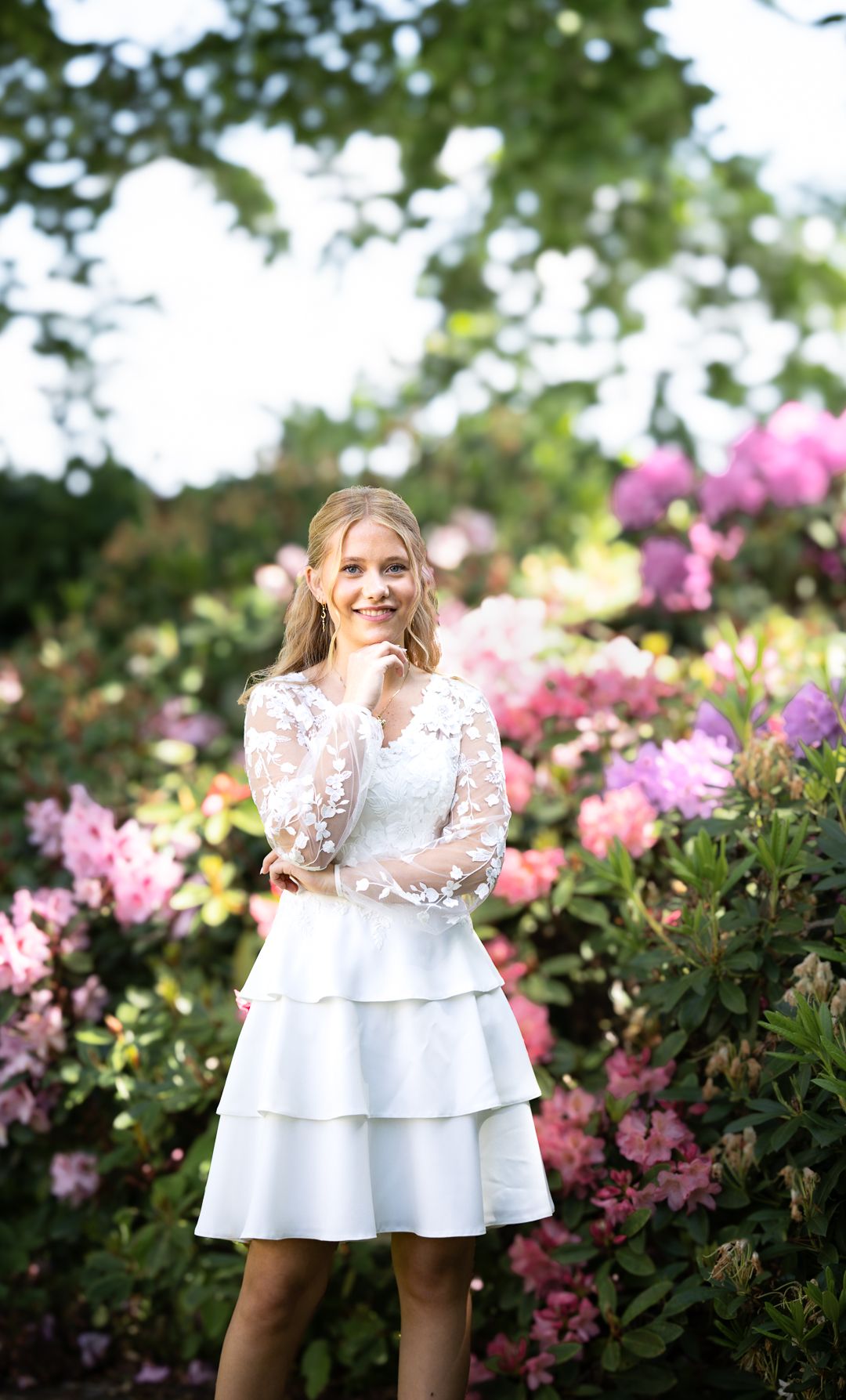Blonde teenage girl in white tiered lace dress smiling among colorful blooming flowers.