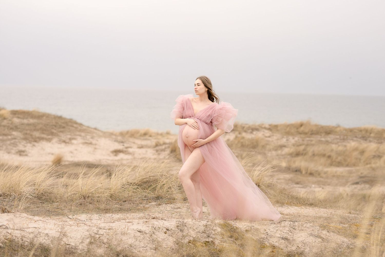 Pregnant woman in flowing pink tulle gown posing gracefully on sandy coastal dunes.