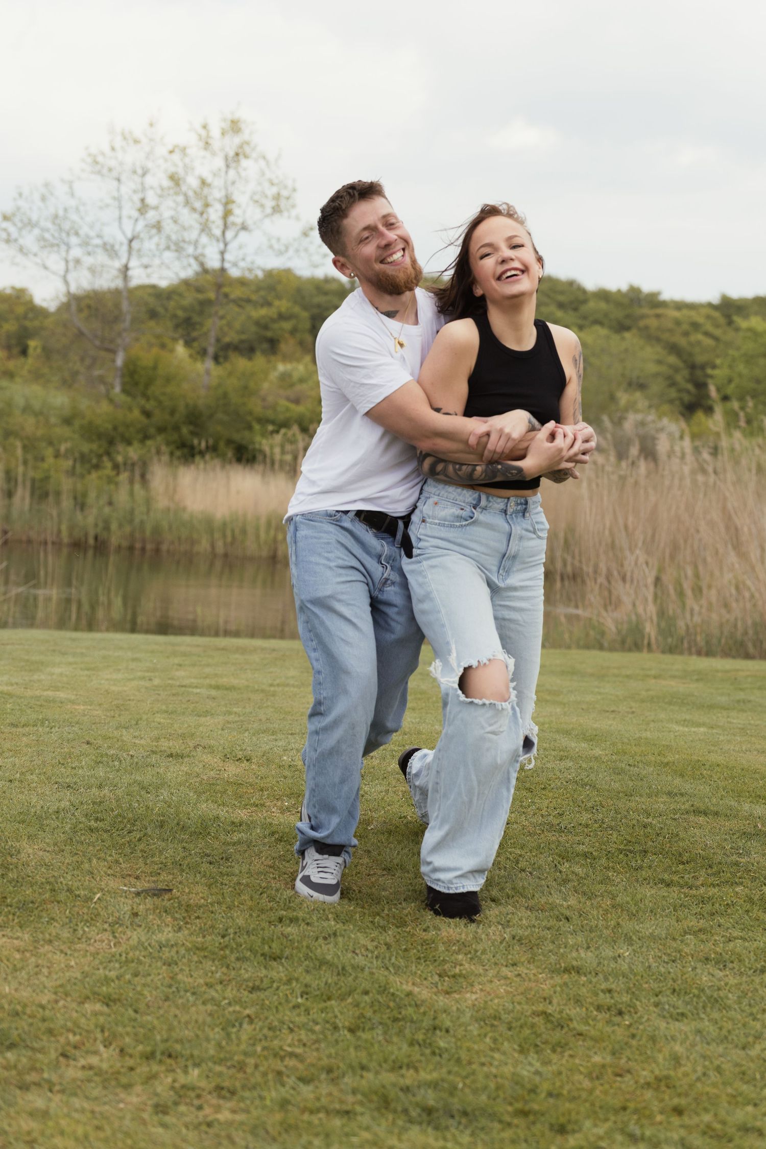 Joyful couple laughing and playing together on a grassy lawn beside a peaceful lake.