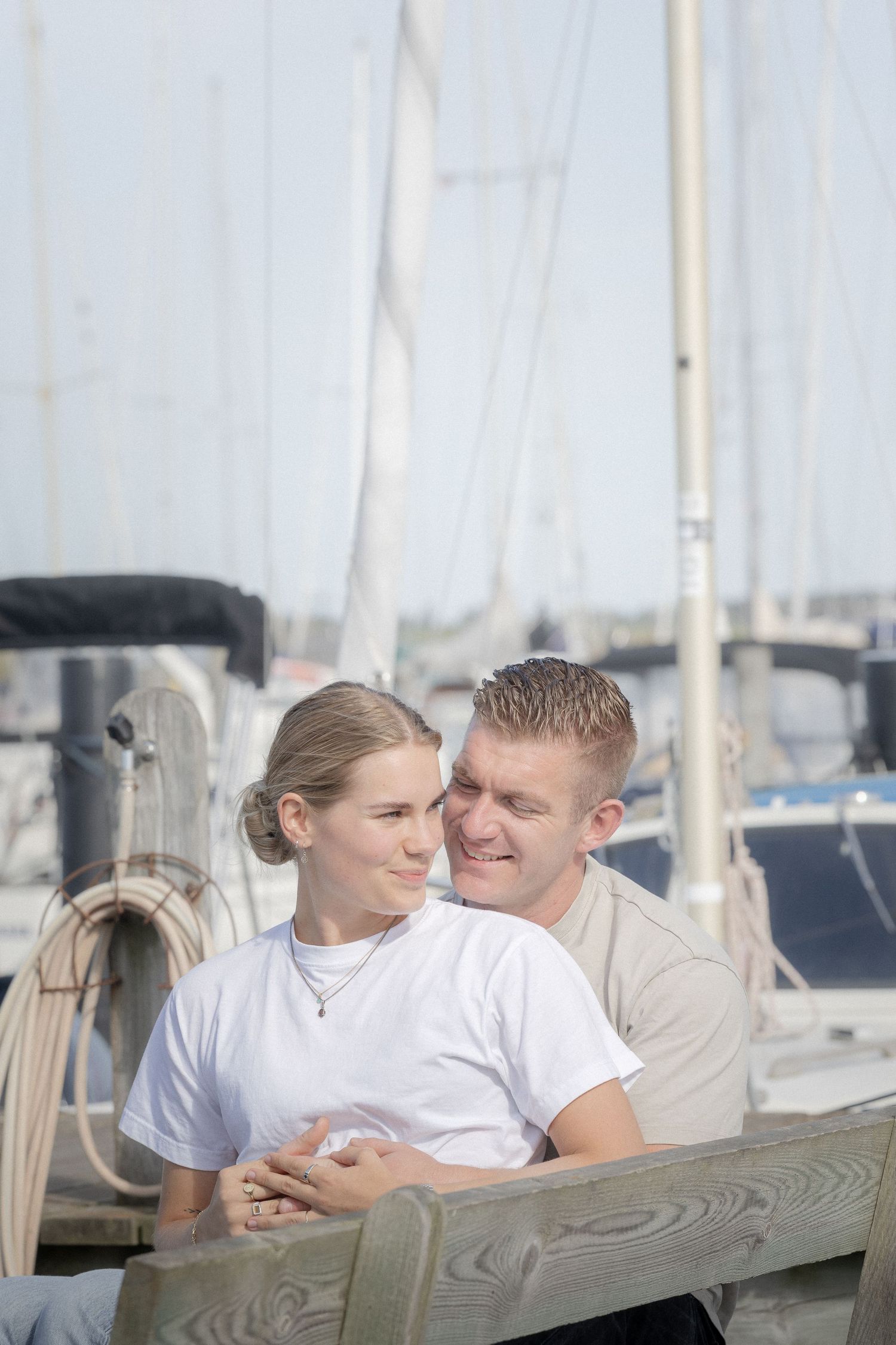 A young couple sits closely together on a wooden bench at a marina, smiling and embracing near sailboats.