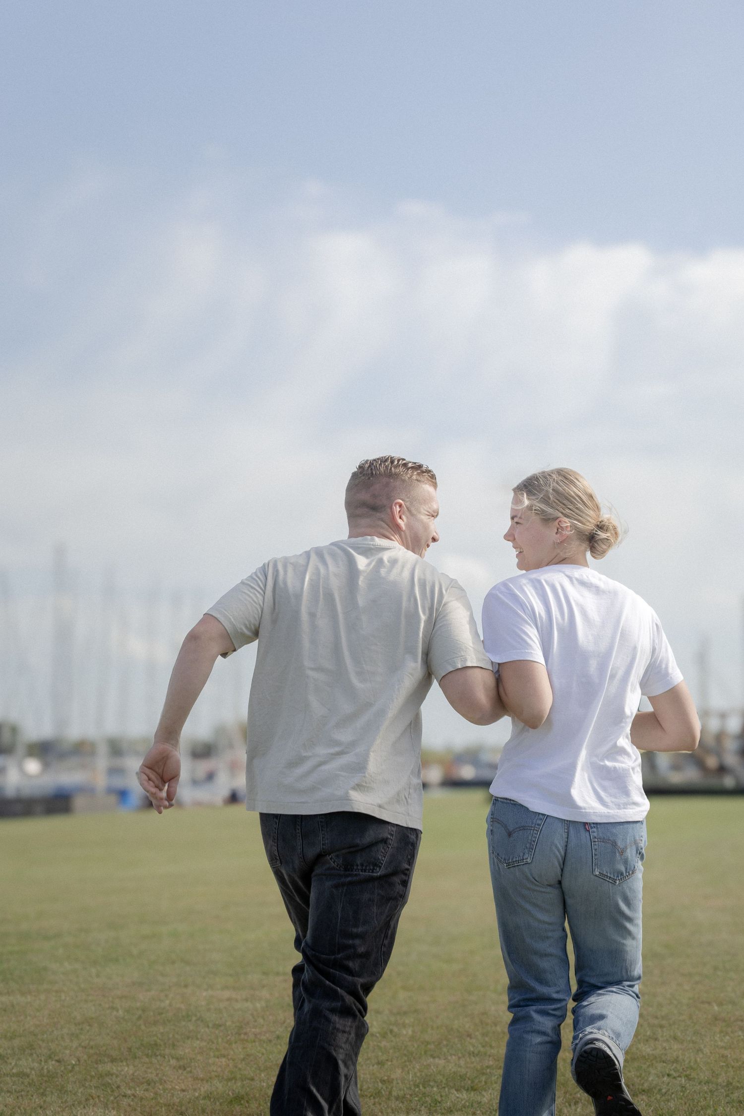 A couple walks across a green field holding hands, smiling at each other with sailboats visible in the distance.