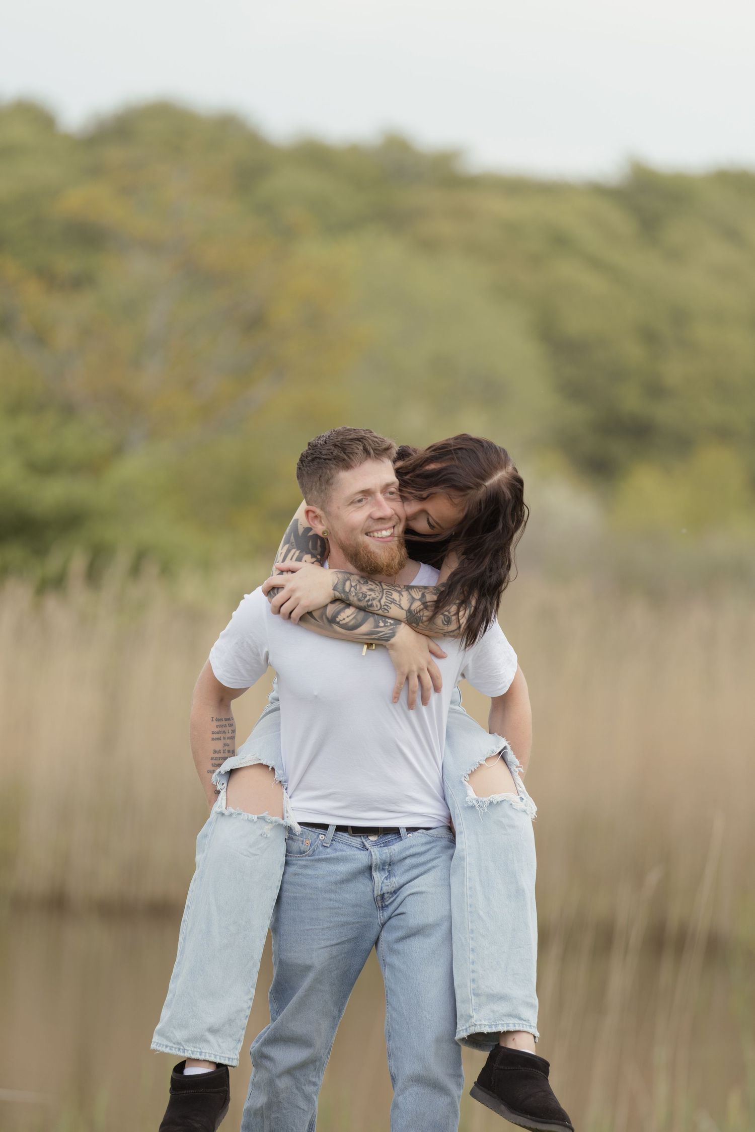 Happy man giving tattooed woman a piggyback ride near a serene lake with tall reeds.