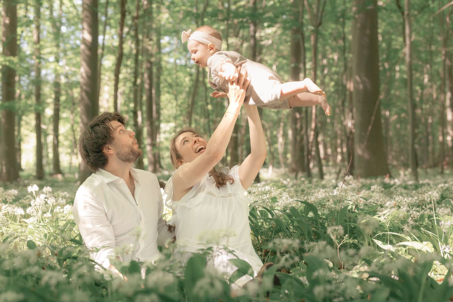 Happy parents lifting smiling baby in sunlit forest clearing with white wildflowers.