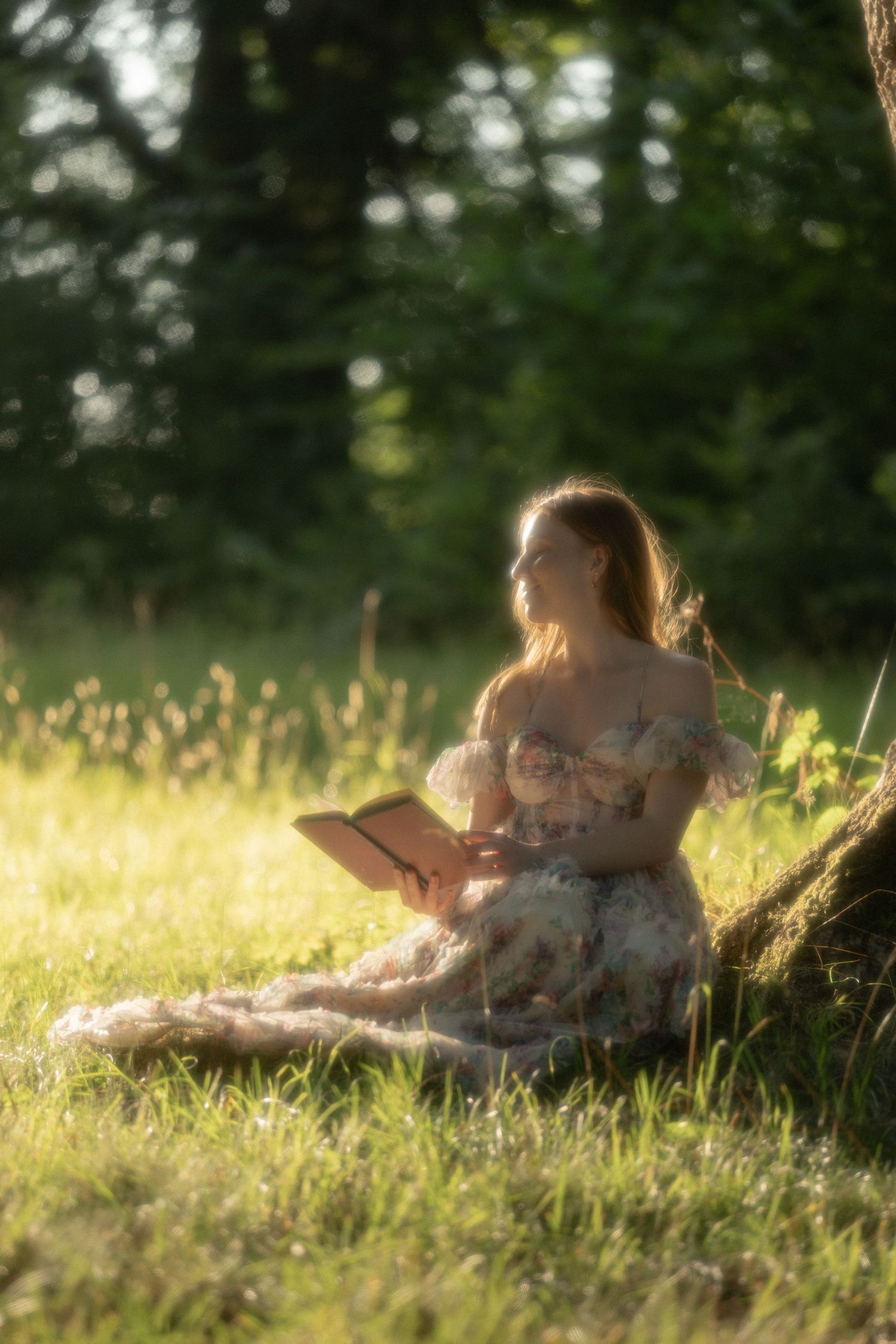 Young woman in floral dress reading a book while sitting against a tree in a sunlit meadow.
