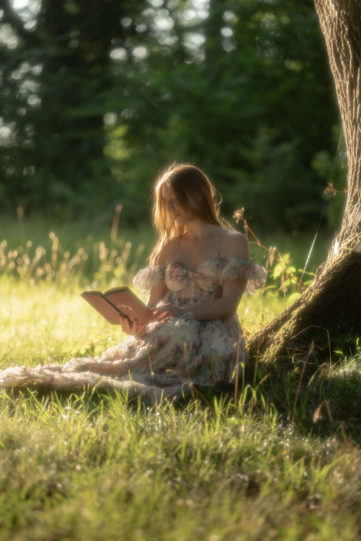 A young woman in a floral dress reads a book while sitting against a tree in a sunlit meadow.