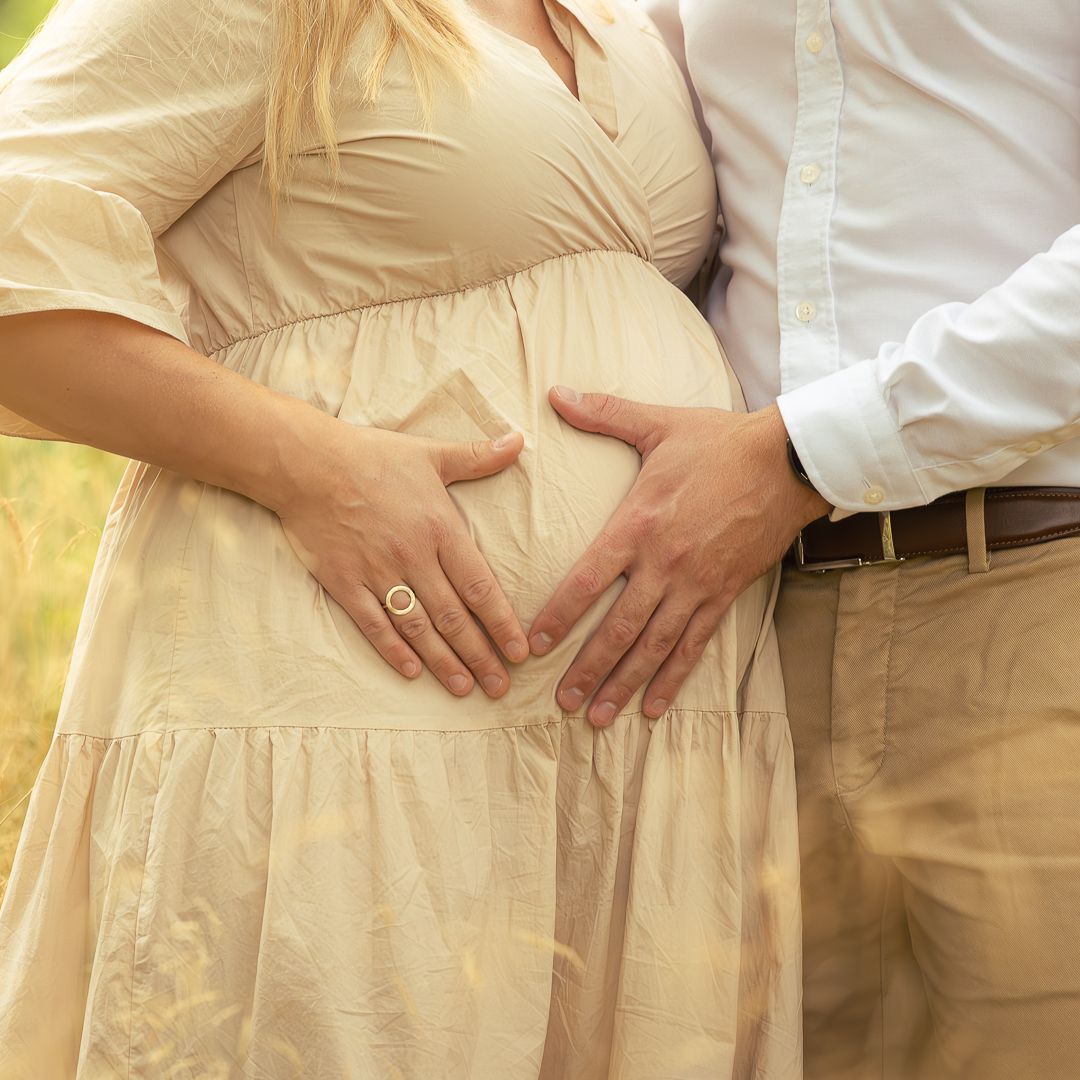 Pregnant woman in beige dress with partner forming heart shape with hands on baby bump outdoors.