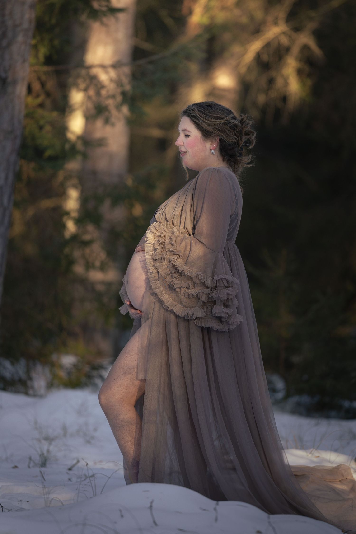 Pregnant woman in sheer taupe gown poses outdoors in snowy winter forest during golden hour maternity photoshoot.