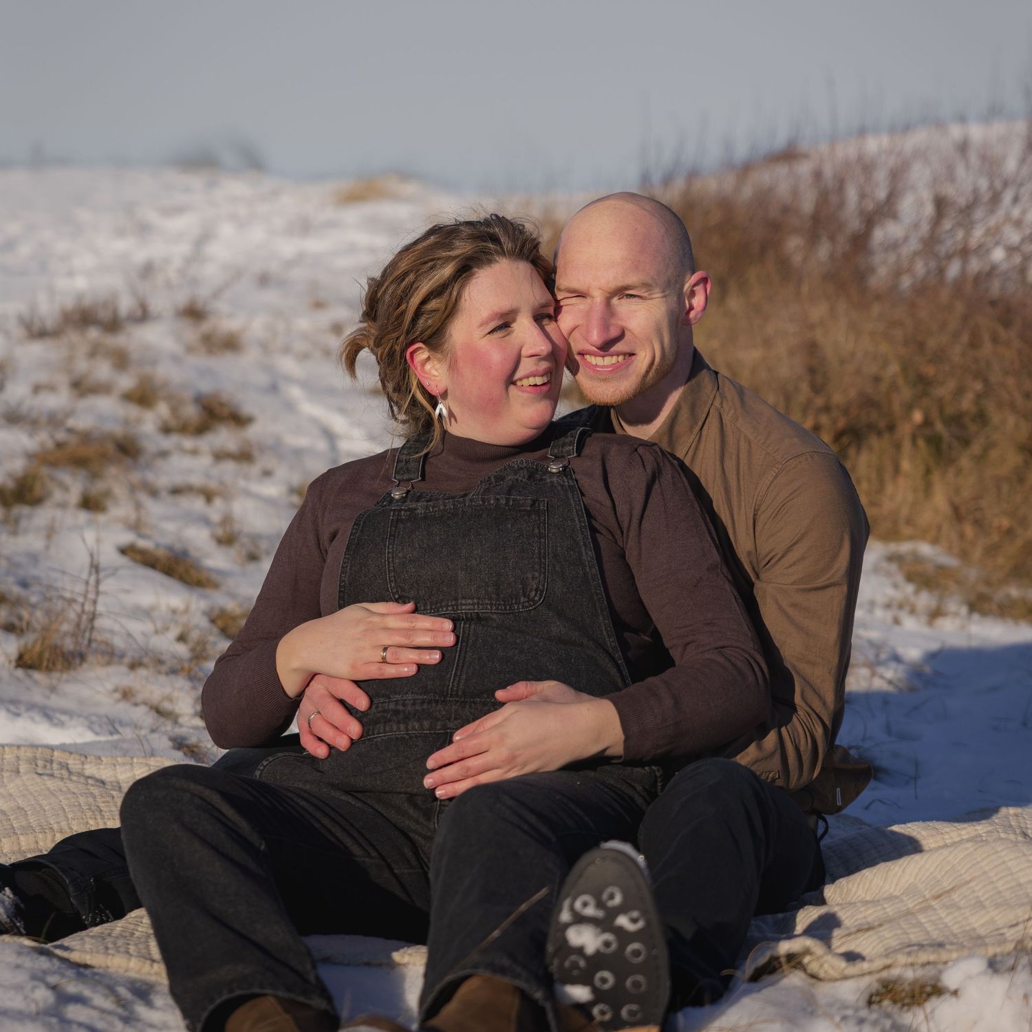 Happy expecting couple sitting together in snowy winter landscape, man embracing pregnant partner warmly.