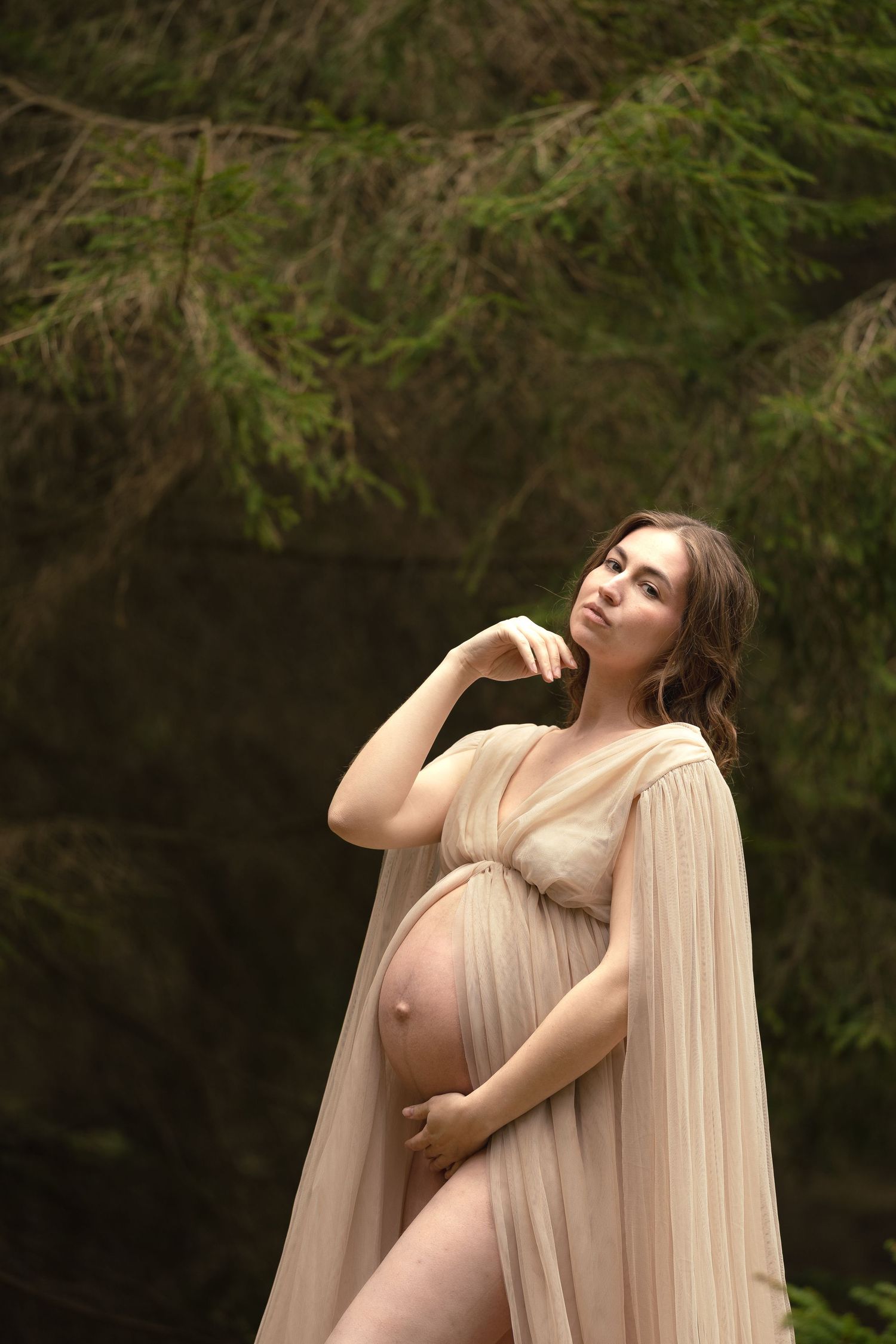 Elegant pregnant woman in draped beige sheer gown gazing upward surrounded by lush green trees.