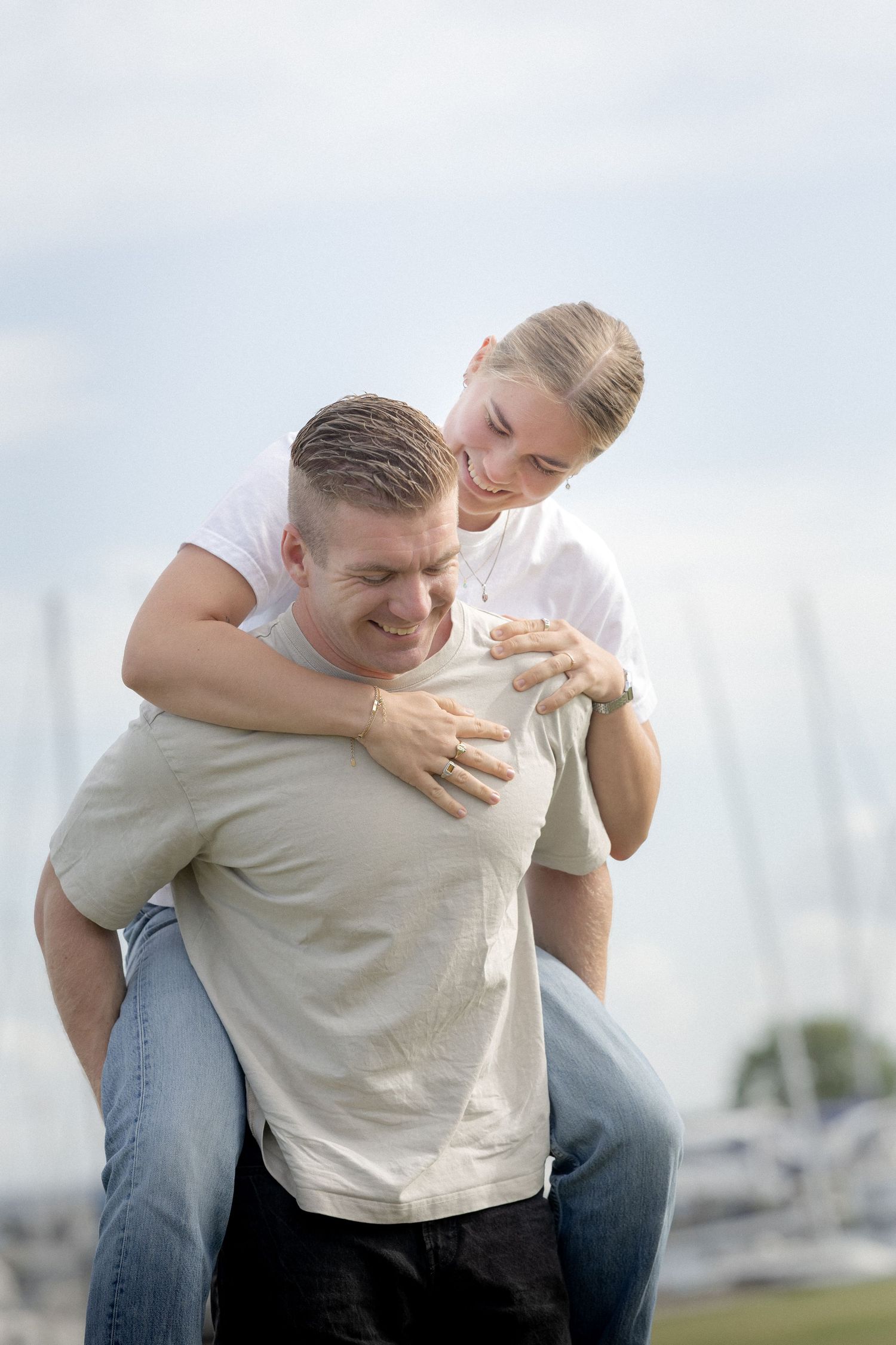 Man giving woman a playful piggyback ride near a marina with sailboats in background.