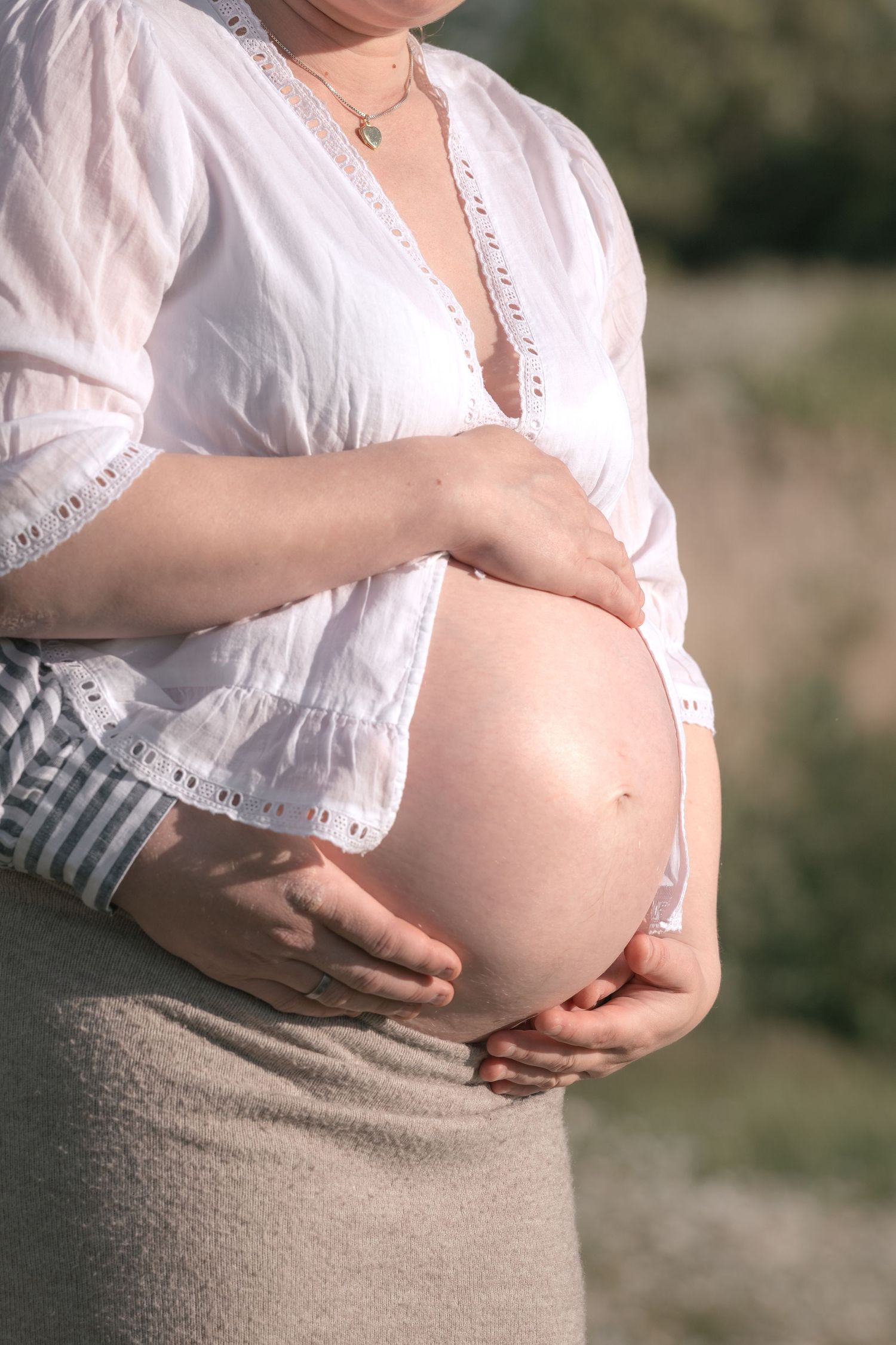 Close-up of pregnant woman in white blouse and beige skirt cradling her bare baby bump outdoors.
