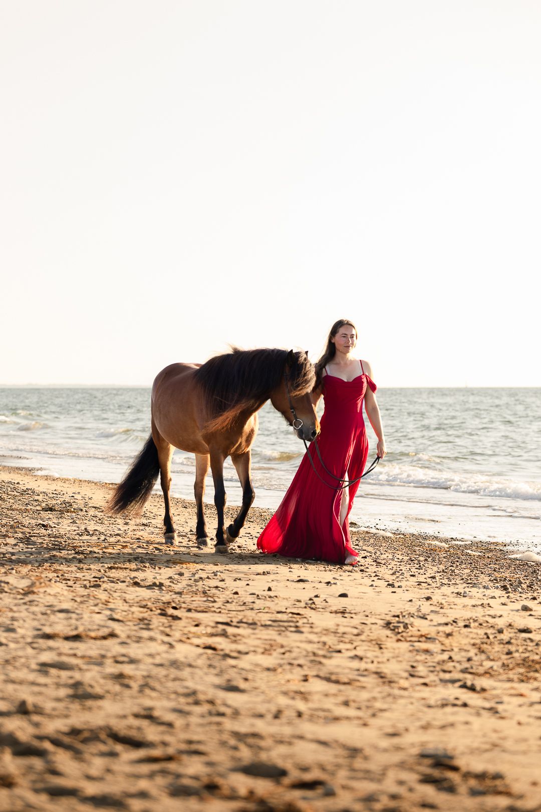 Elegant woman in red gown walking with brown horse along scenic beach at sunset.